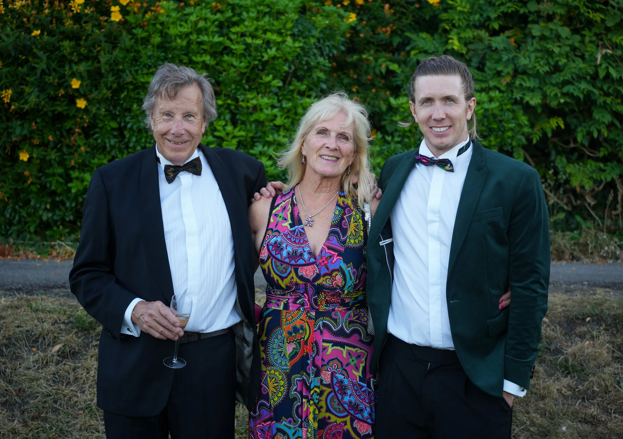 A group of three people dressed formally, standing outdoors with green bushes in the background. The older man on the left is wearing a black tuxedo with a bow tie and holding a glass of wine. The woman in the center is wearing a colorful, patterned 