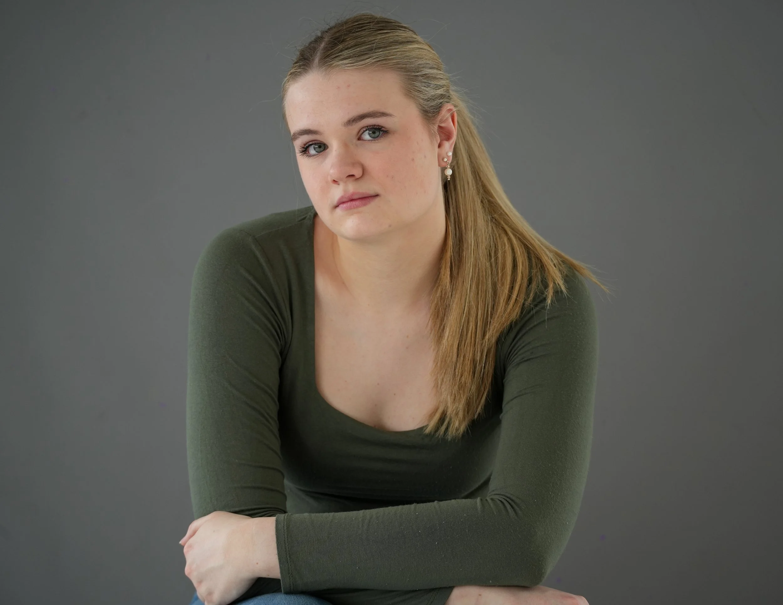 A young woman with blonde hair tied back, wearing a dark green top, sitting with her arms crossed against a plain gray background.