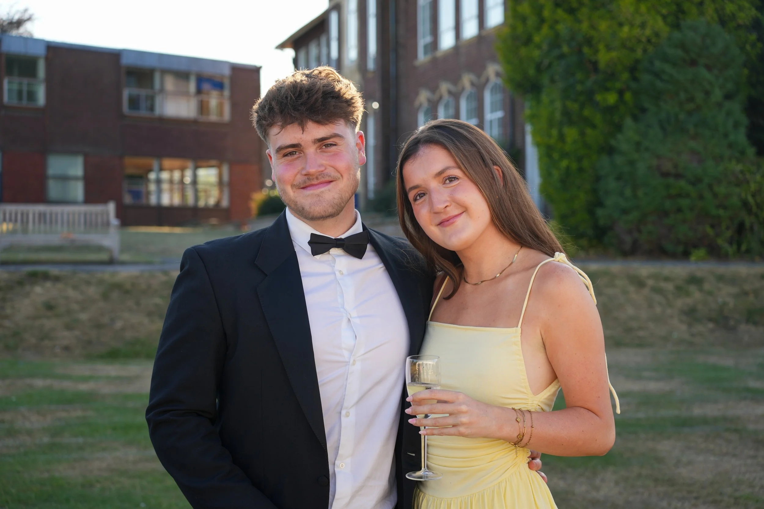 A young man in a tuxedo and a young woman in a yellow dress smiling while holding a glass of champagne, standing outdoors in front of brick buildings and greenery.