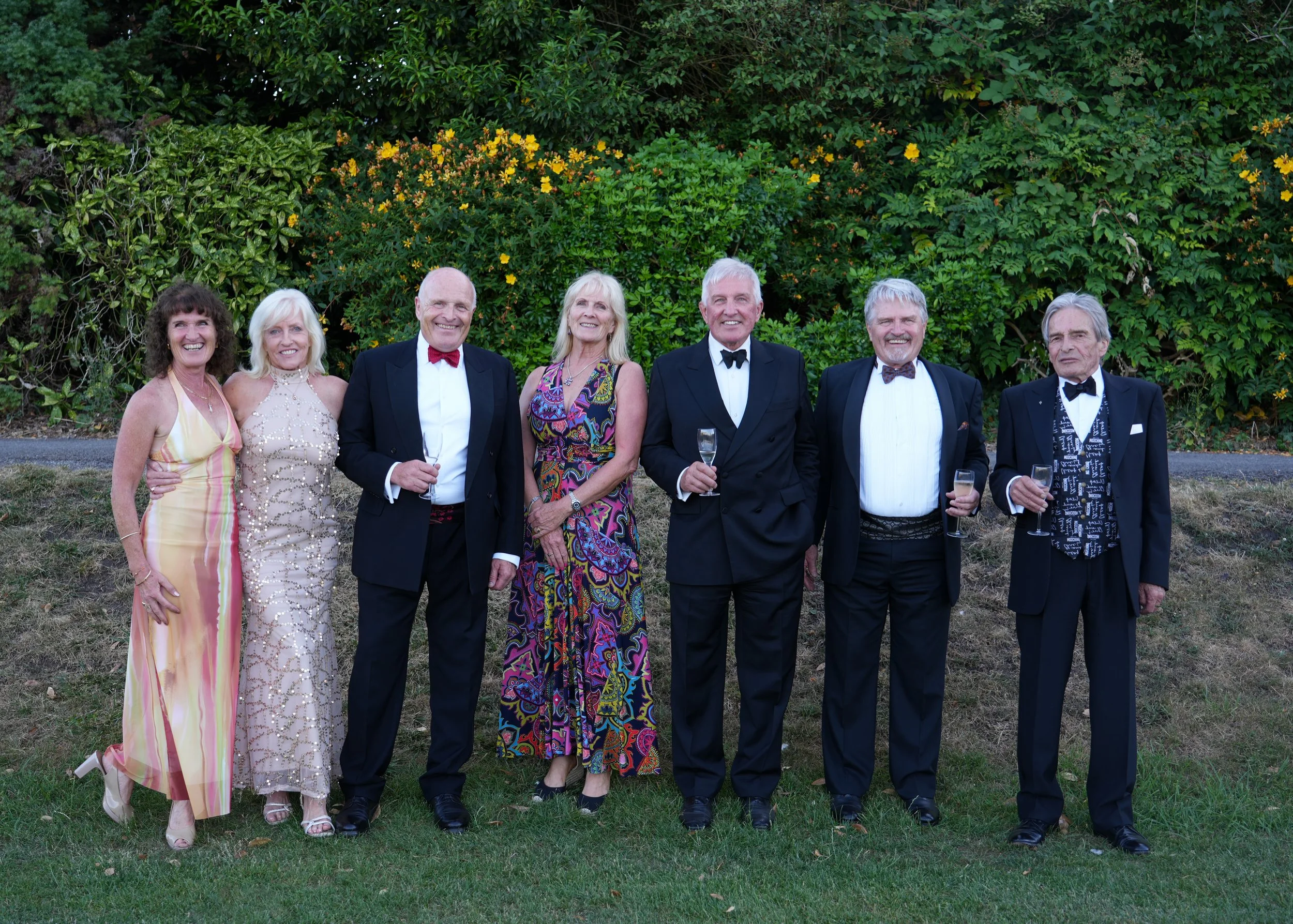 Group of seven older adults in formal attire standing on grass with a green, leafy background, smiling and holding champagne glasses, at an outdoor event.