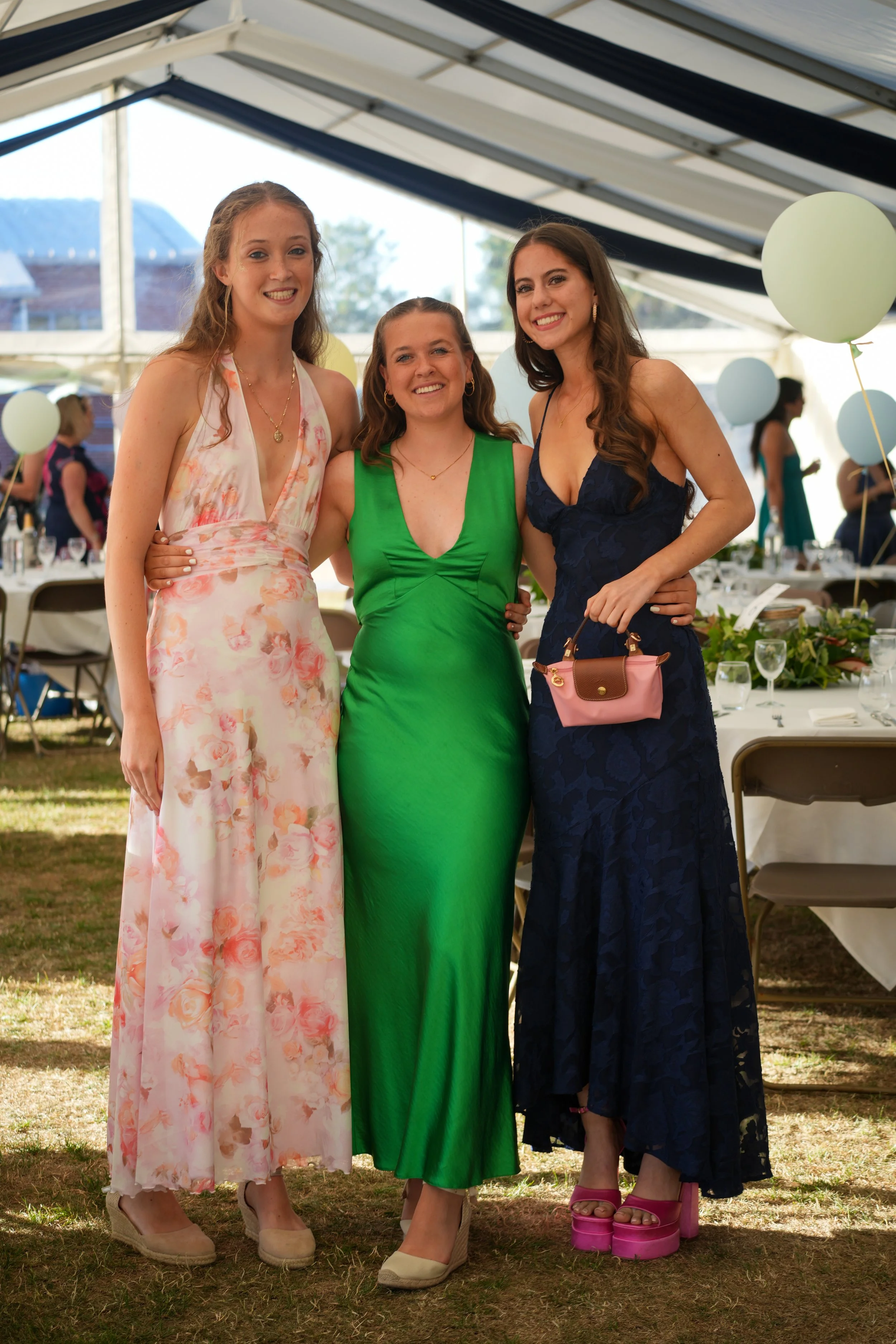 Three women standing together at a party under a tent, smiling and posing for a photo. The woman on the left is wearing a floral dress, the woman in the center is in a bright green dress, and the woman on the right is in a dark blue lace dress with pink platform shoes, holding a small purse.