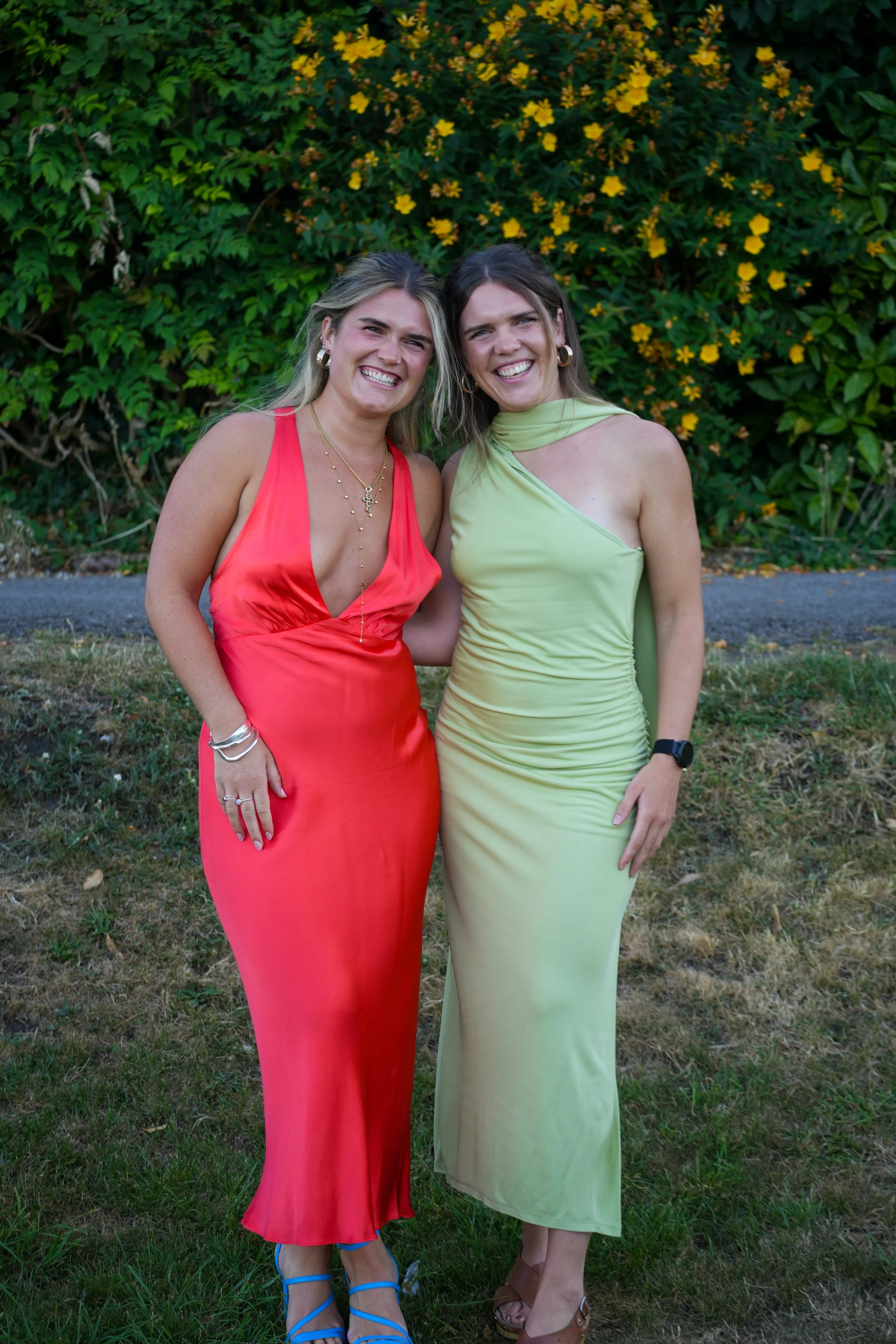 Two women standing outdoors in front of green shrubbery with yellow flowers, smiling. The woman on the left is wearing a sleeveless red dress with a deep V-neck and blue strappy heels, while the woman on the right is wearing a light green sleeveless dress with a matching fabric drape on her shoulder and tan sandals.