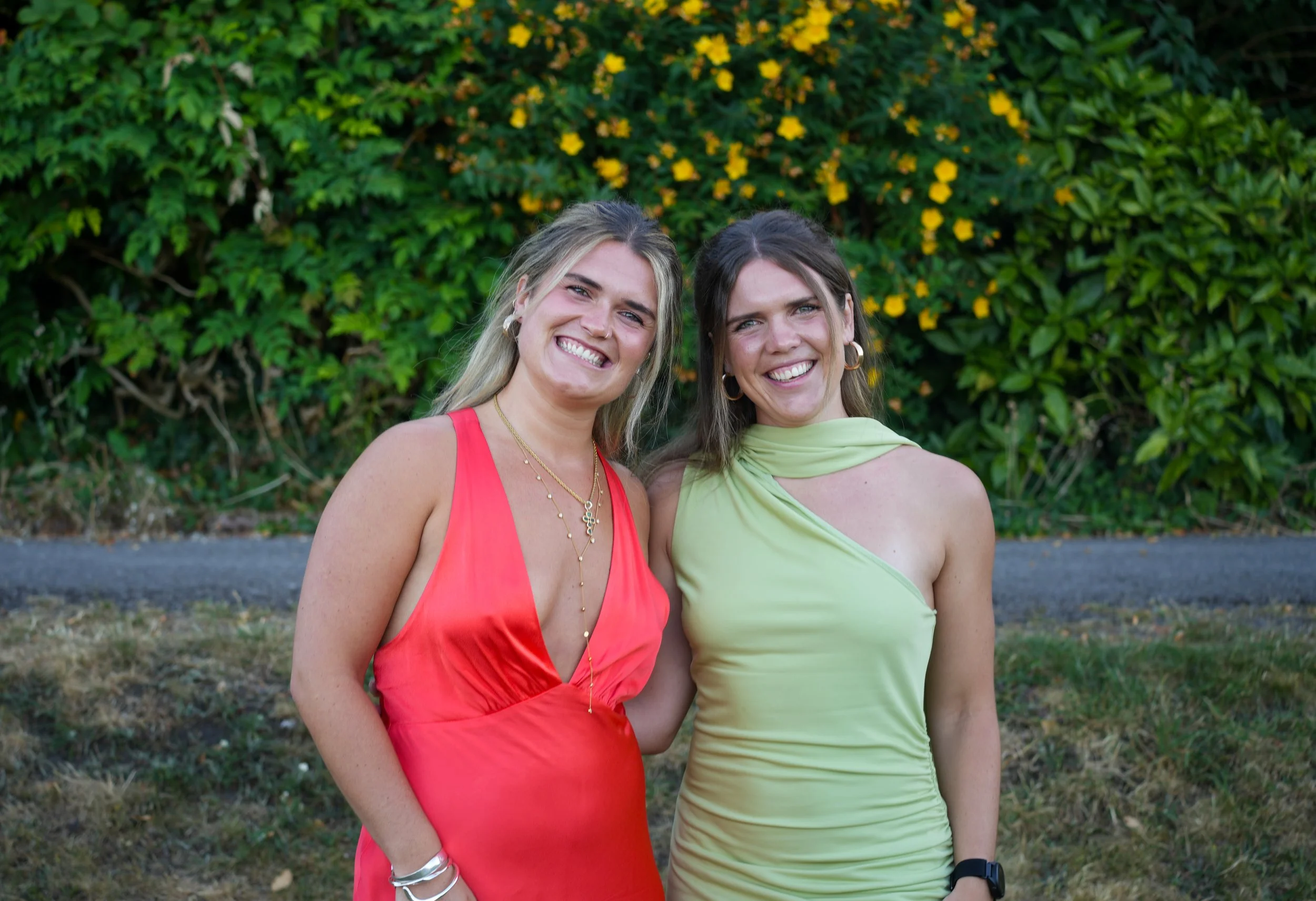Two smiling women standing outdoors in front of green foliage, wearing colorful dresses, one in coral and the other in lime green.