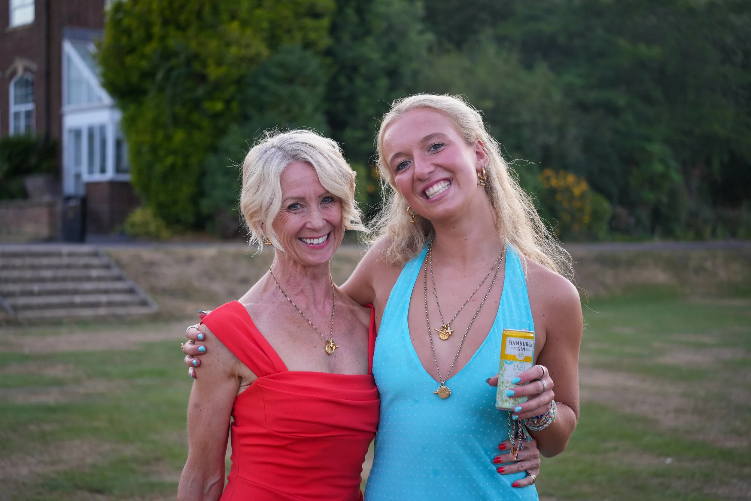 A woman in a red dress and an older woman in a blue dress smiling and embracing outdoors.