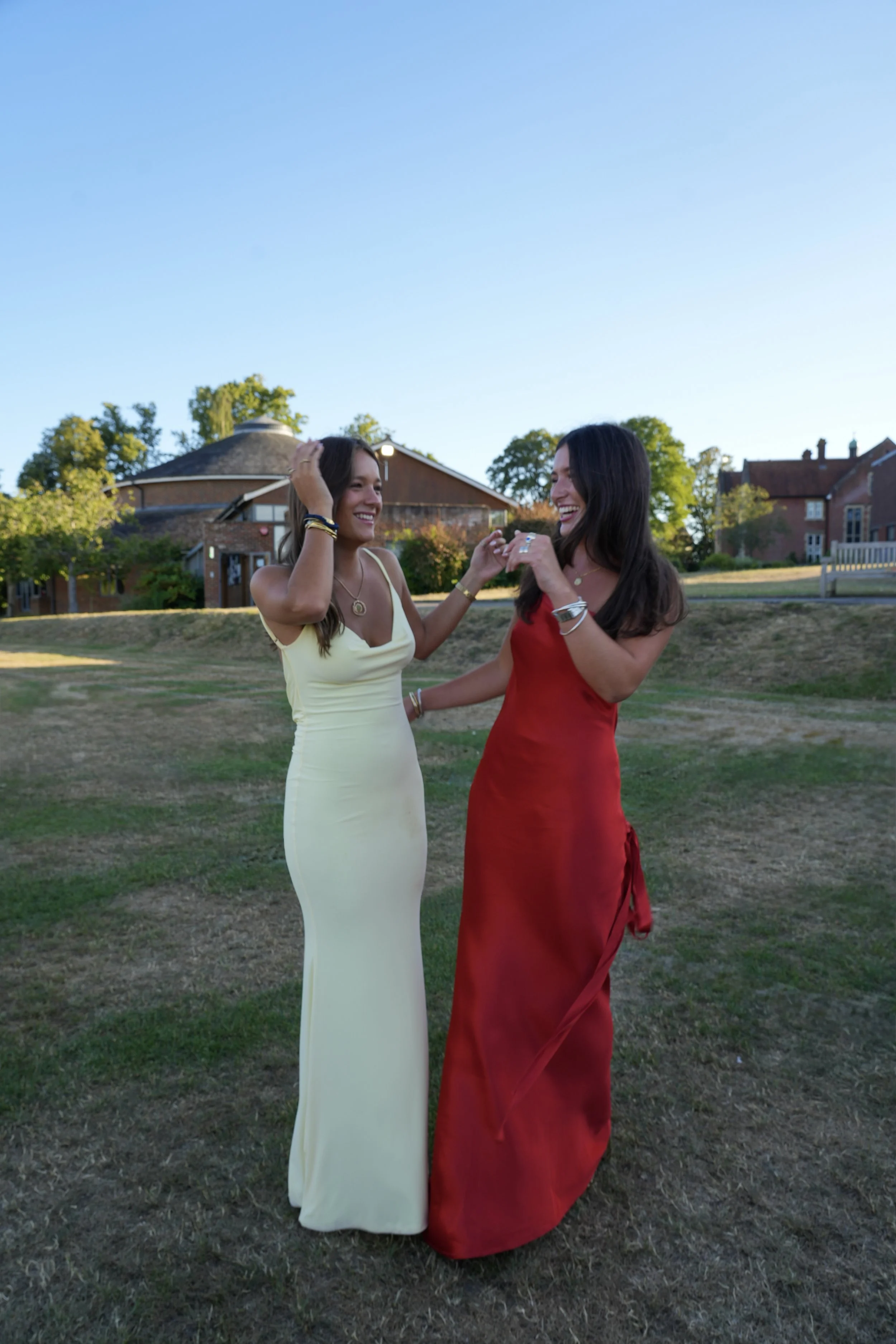 Two women in formal dresses laughing and talking outdoors on a grassy area with buildings and trees in the background during daytime.