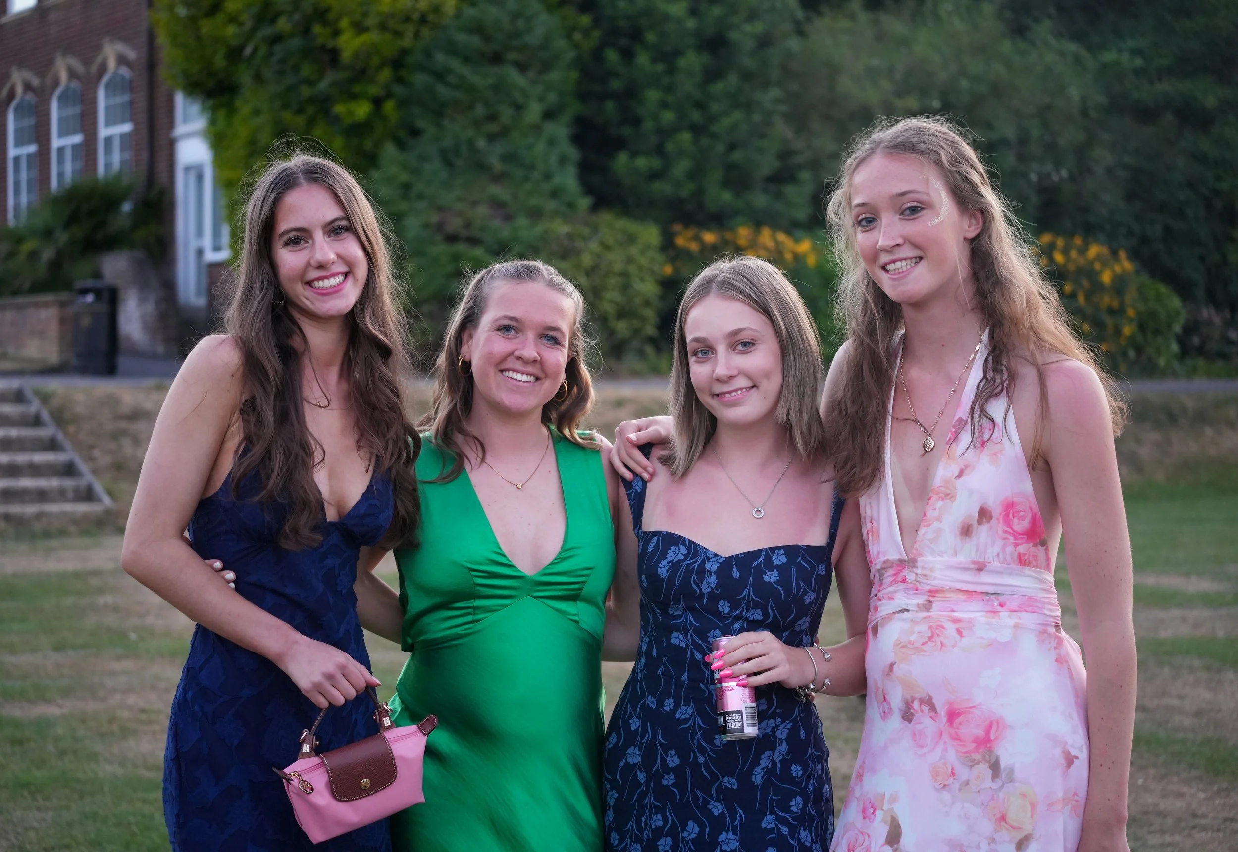 Group of five young women smiling outdoors, some holding drinks, standing on a grassy area with trees and a brick building in the background.