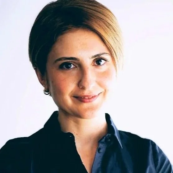 A woman with short, light brown hair in a bob style, wearing a navy blue collared shirt, smiling against a plain white background.