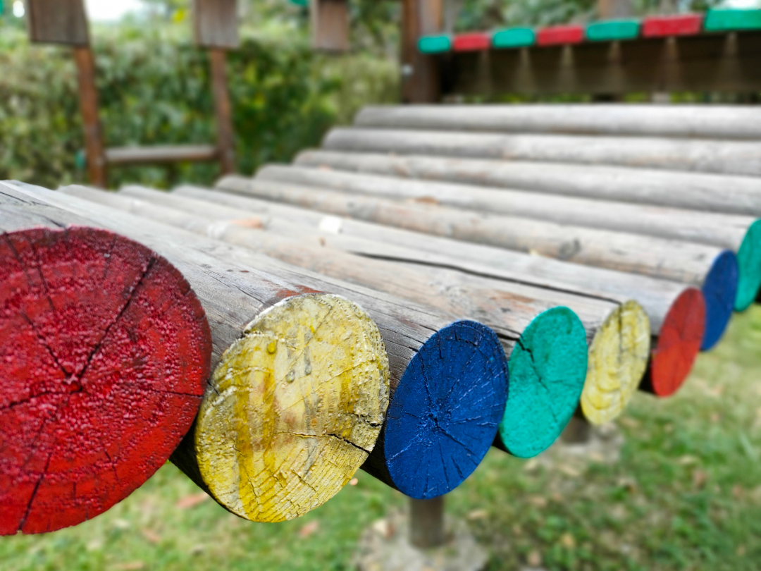Barra de madera con partes cortadas y pintadas de diferentes colores en un parque o zona de juegos al aire libre.