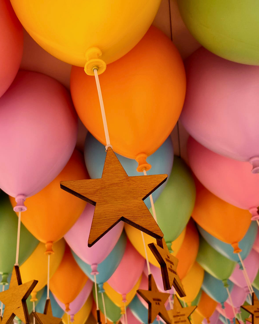 Globos de colores rosa, naranja, verde y azul decorados con estrellas de madera colgadas con hilos blancos.