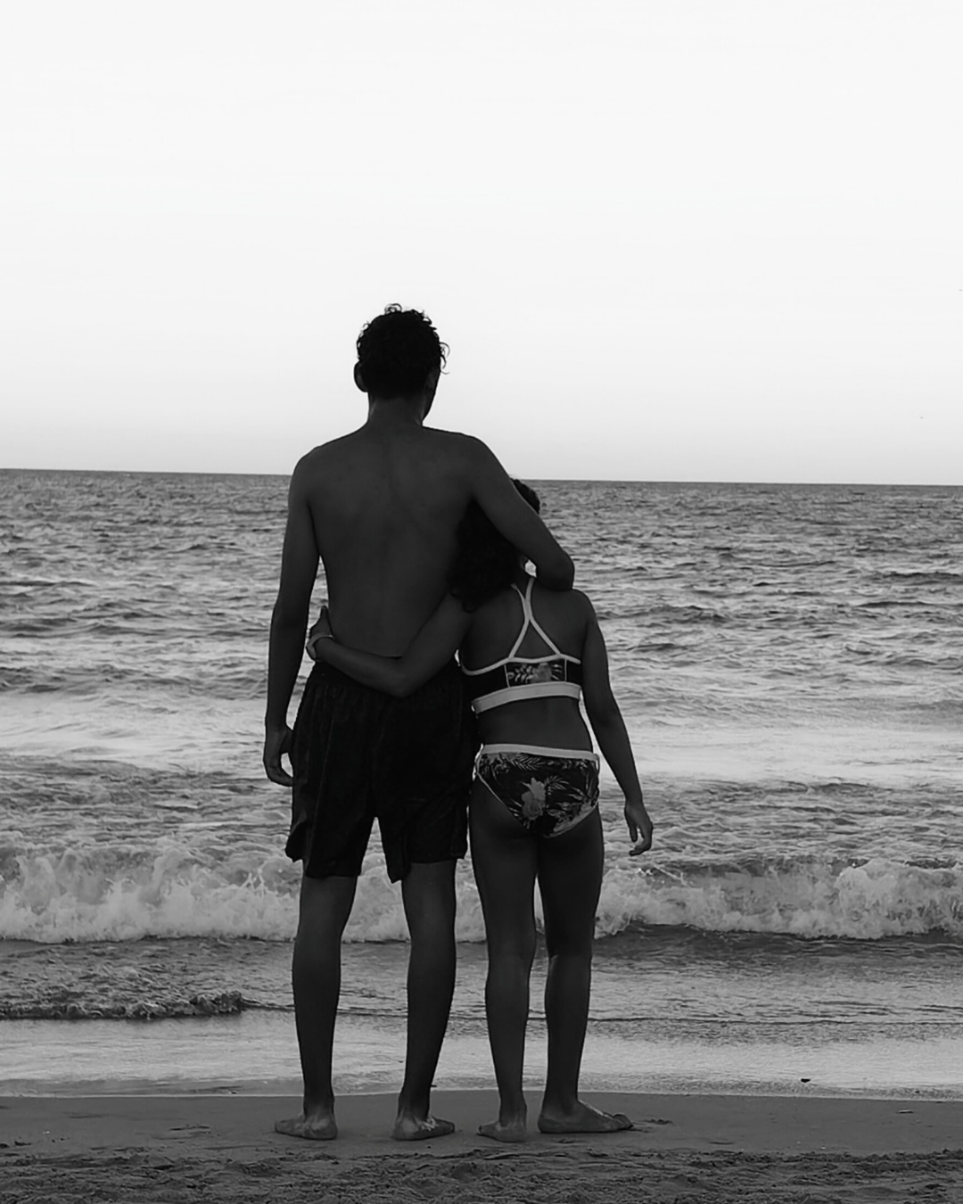 Una pareja joven, el hombre sin camisa y la mujer con traje de baño, se abraza en la playa mirando el mar en un atardecer o amanecer.