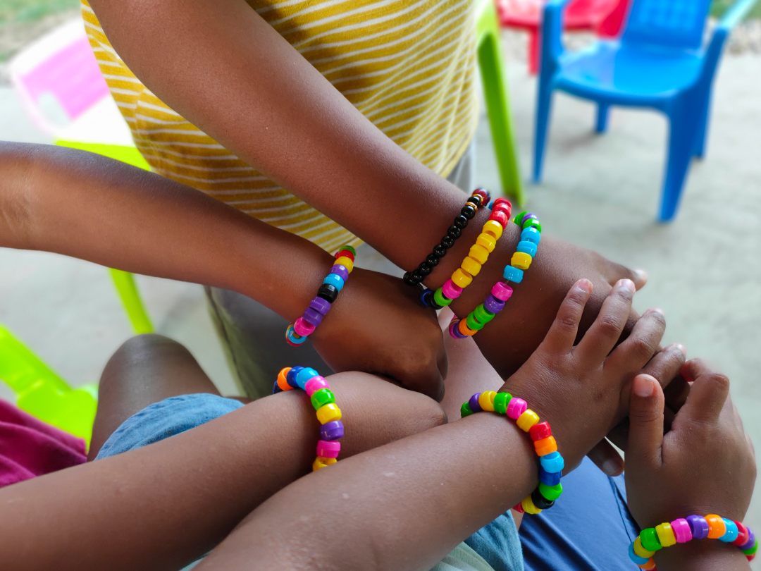Grupo de niños con pulseras de colores, mostrando la unión de sus manos, en un ambiente exterior con sillas de plástico de varios colores.