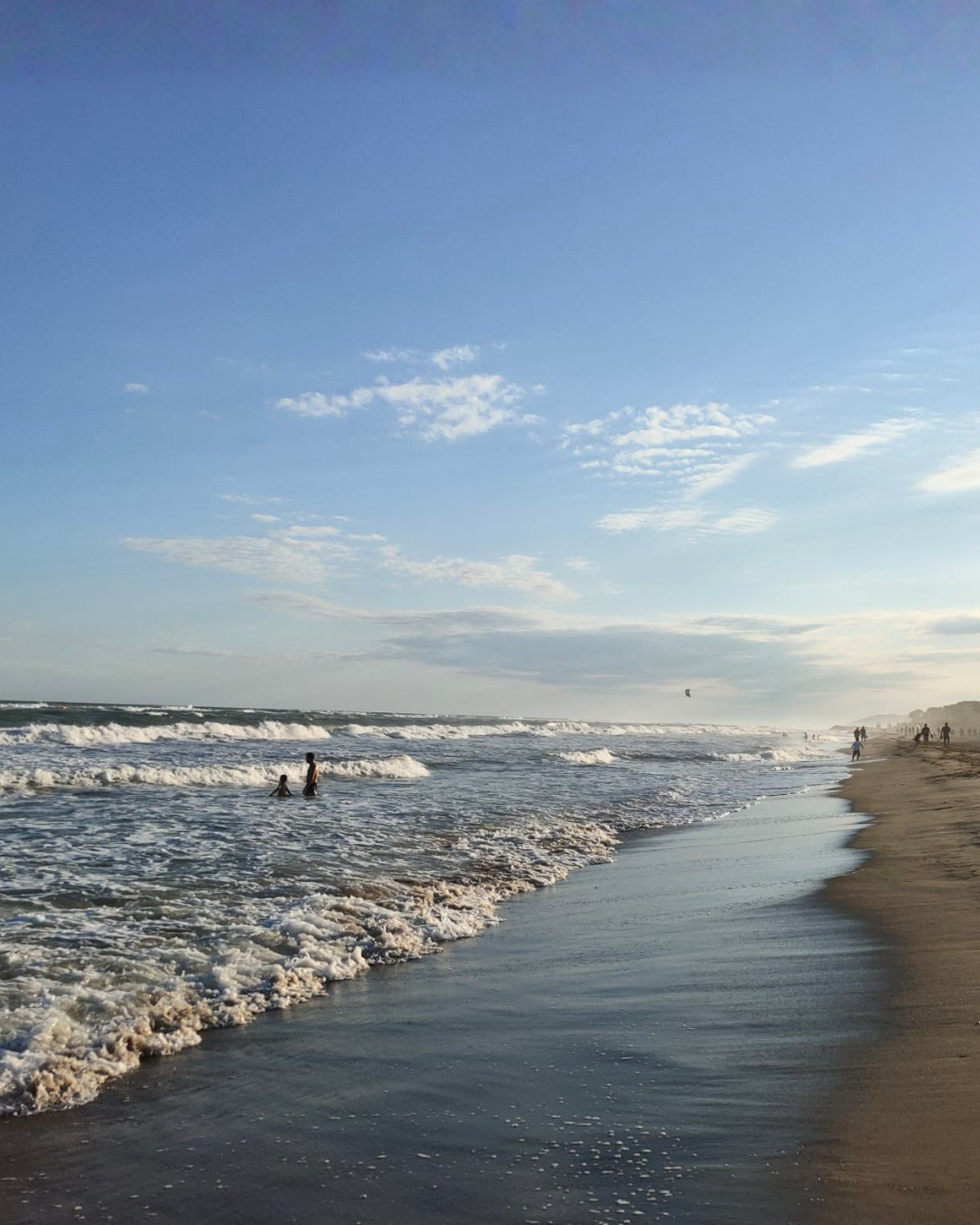 Vista de una playa con arena dorada, olas rompiendo en la orilla, varias personas disfrutando del mar, cielo azul con algunas nubes y un parapente en el aire en la distancia.