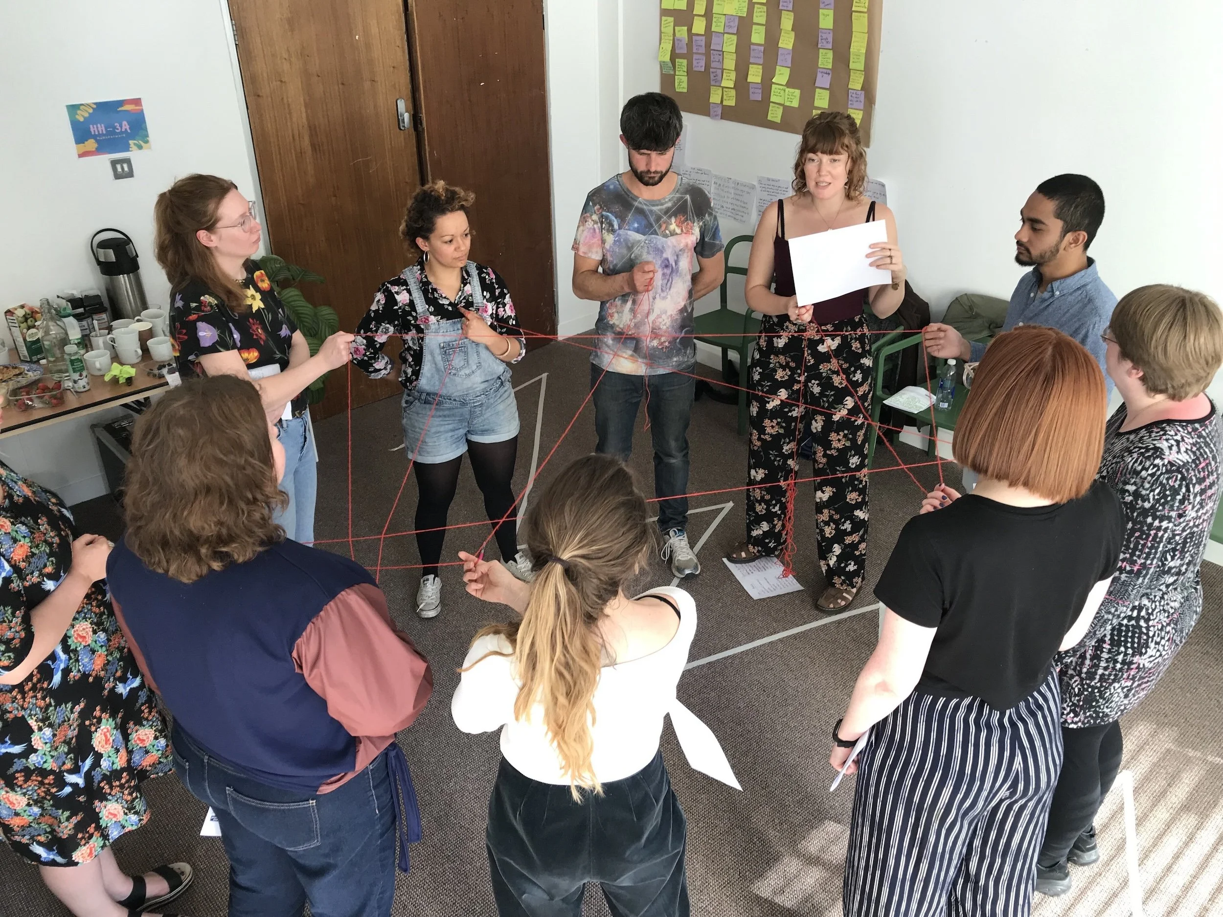 Group of diverse people standing in a circle, engaged in team-building activity with red string, in a room with white walls, a bulletin board with colorful notes, a table with cups and snacks, and a closed wooden door.