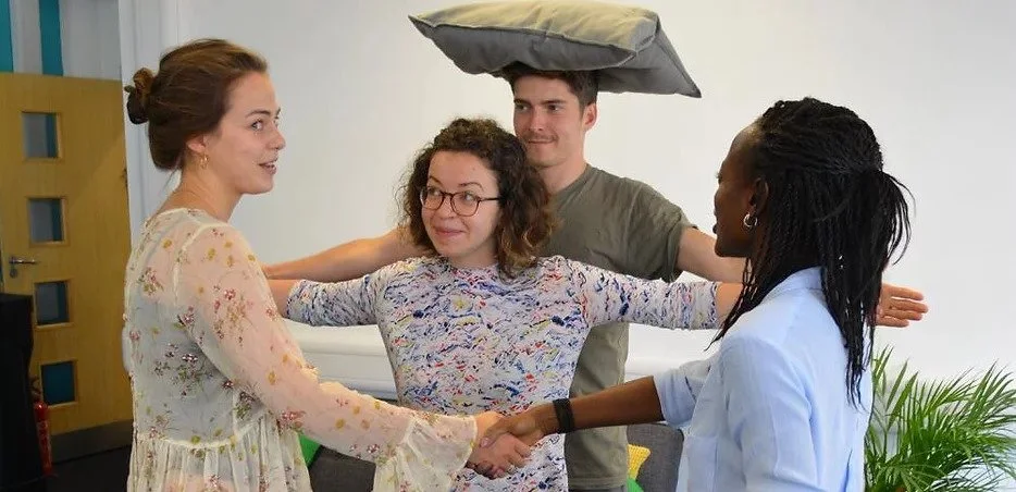 Four people in an office shaking hands and smiling, with one person standing on a raised pillow on another's shoulders, creating a humorous scene.