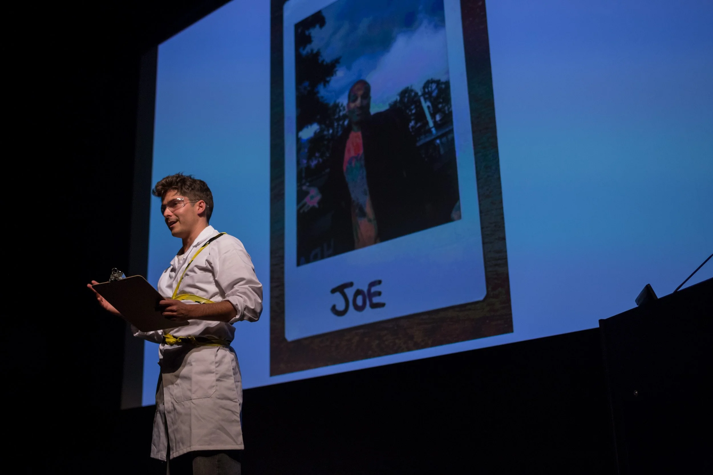 Young man in white shirt holding clipboard giving presentation on stage with large backdrop of a Polaroid photo labeled "Joe" and showing a person outdoors with trees.