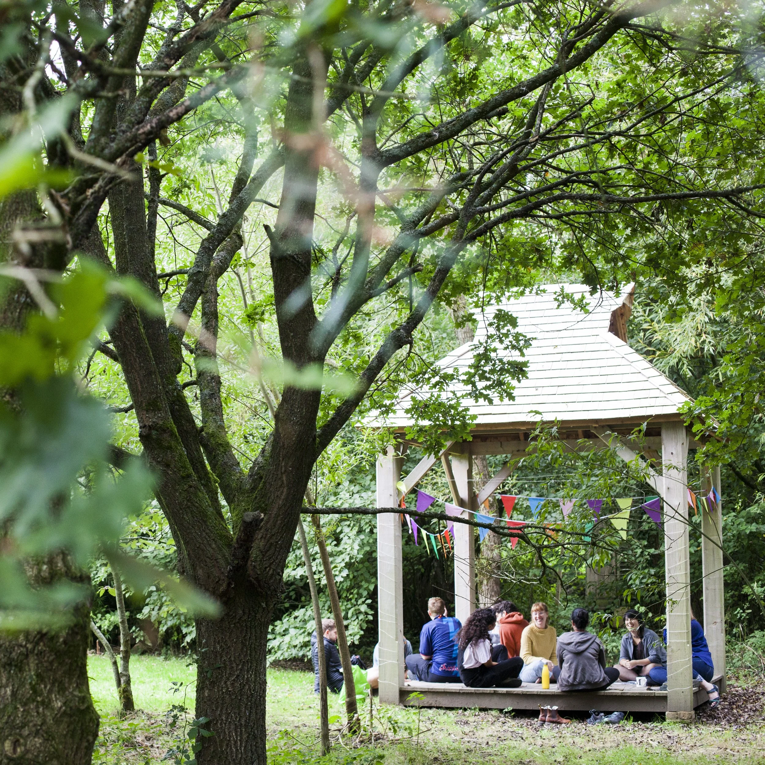 Group of people sitting and chatting inside a wooden gazebo decorated with colorful bunting, situated in a lush green outdoor setting with trees.