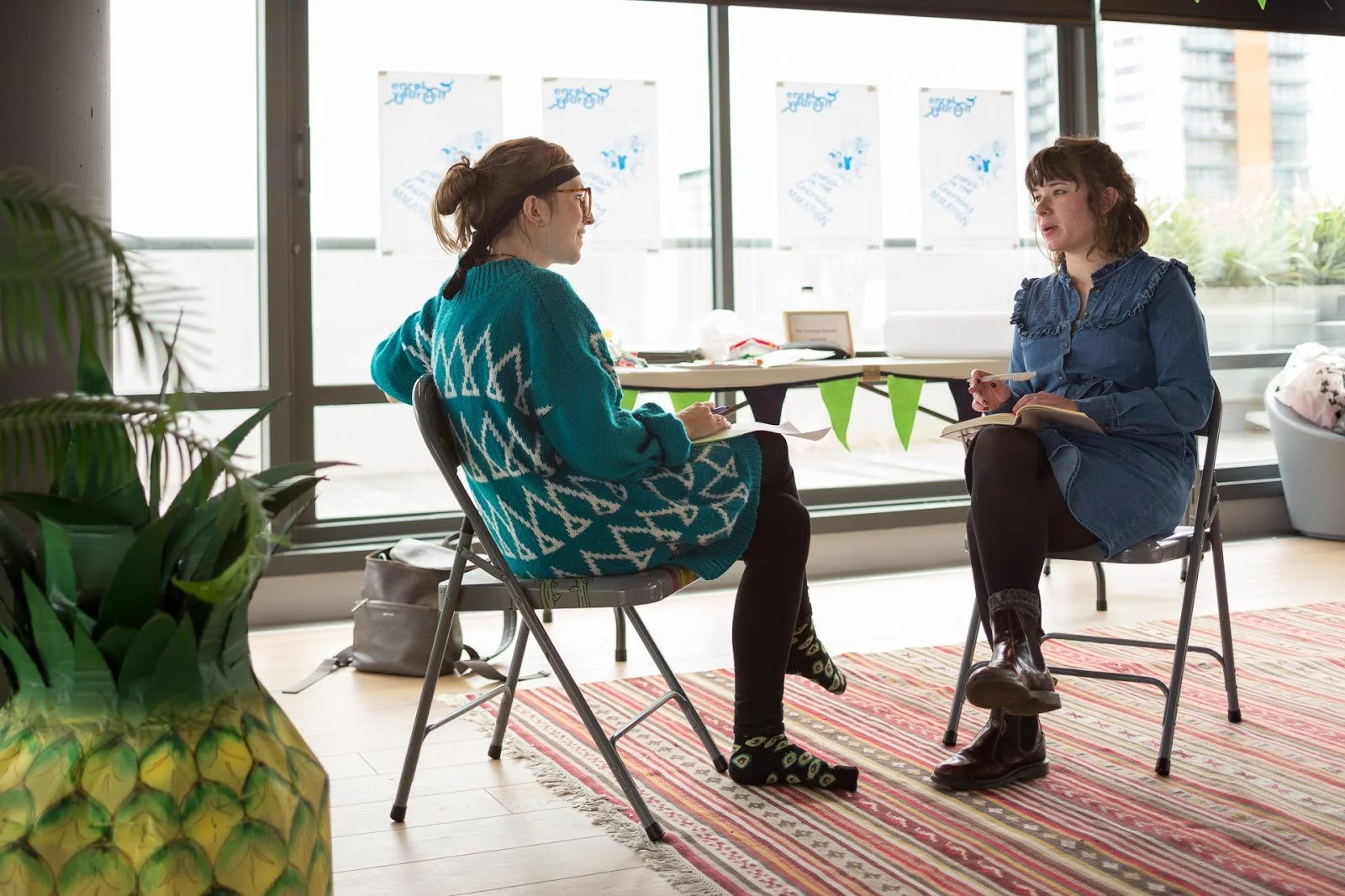 Two women sitting and talking in an indoor space with large windows and urban buildings outside. One woman wears a blue sweater with a white geometric pattern, glasses, and patterned socks. The other woman wears a blue dress with ruffles, black tights, and boots, and is holding a notebook and pen.