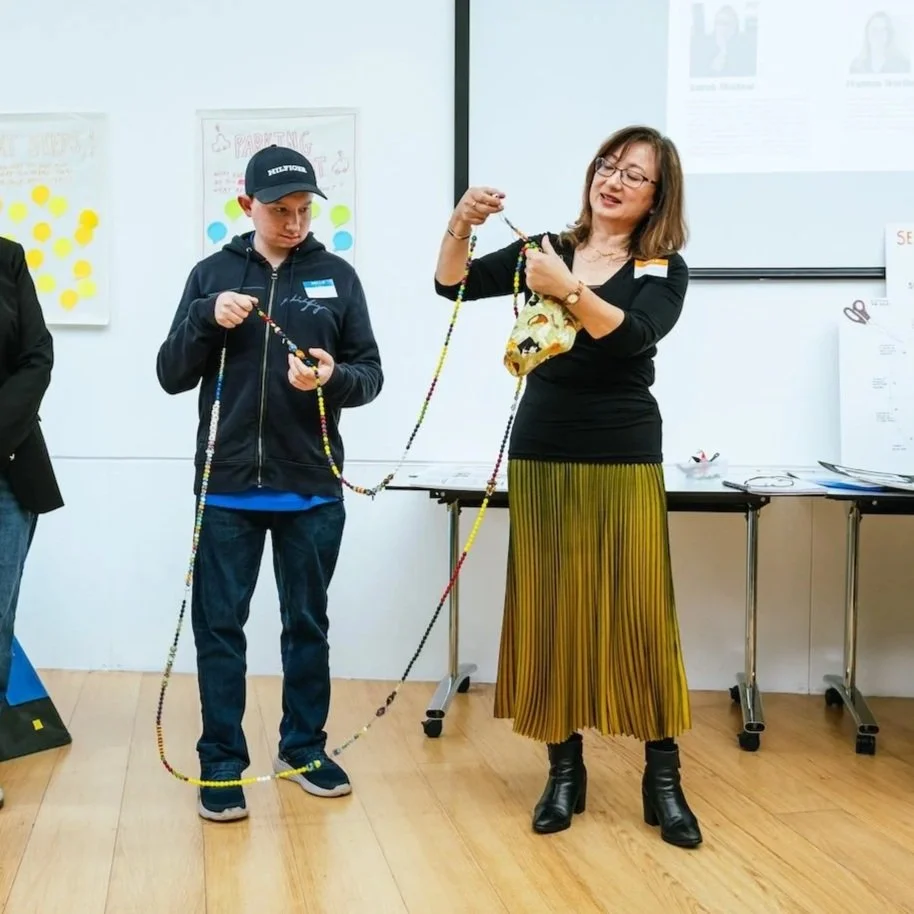 A woman and a boy holding a long beaded necklace together in the Economic Justice Huddles Showcase. Presenting on behalf of the SEND (Special Educational Needs and Disabilities) Seen Huddle. 