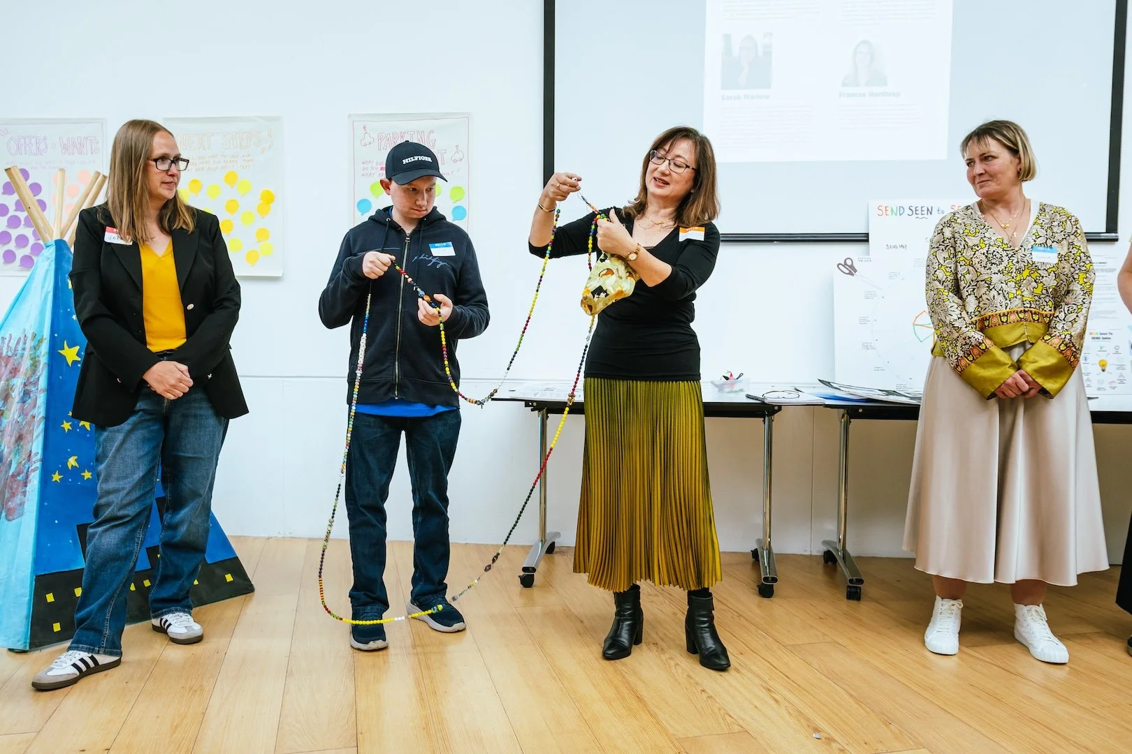 Group of four women and one young man standing in a room, with one woman demonstrating how to make a craft string with beads, while the others observe. There are colorful posters on the wall and a digital screen behind them.
