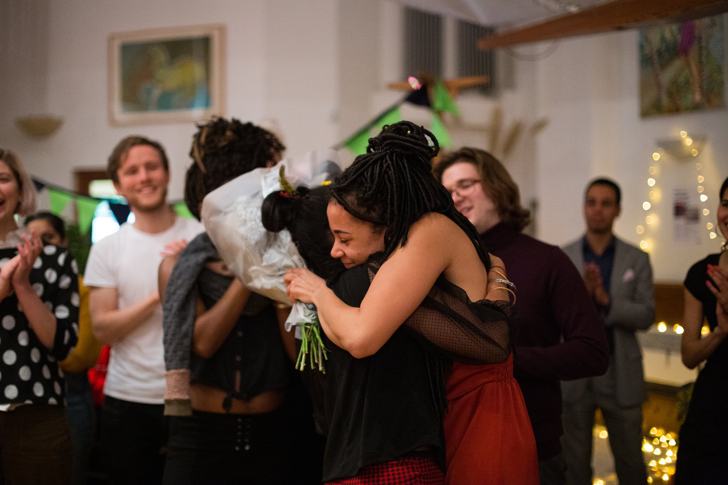 People hugging at a celebration, some clapping and smiling, decorated with string lights and party decorations.