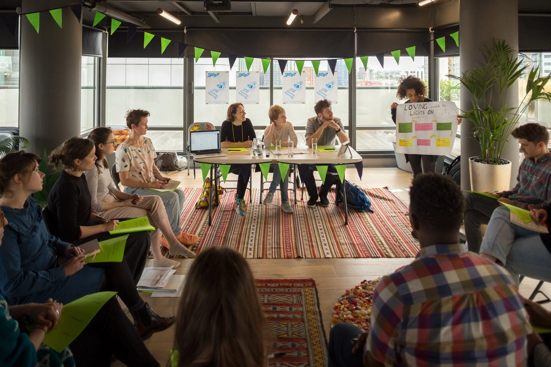 Group of people participating in a meeting or workshop in a modern, well-lit room with large windows and green and purple triangular banners hanging from the ceiling. One person stands presenting a poster to the audience, while others sit around a large table or on chairs, some holding green folders or notebooks. The room has plants and decorative rugs.