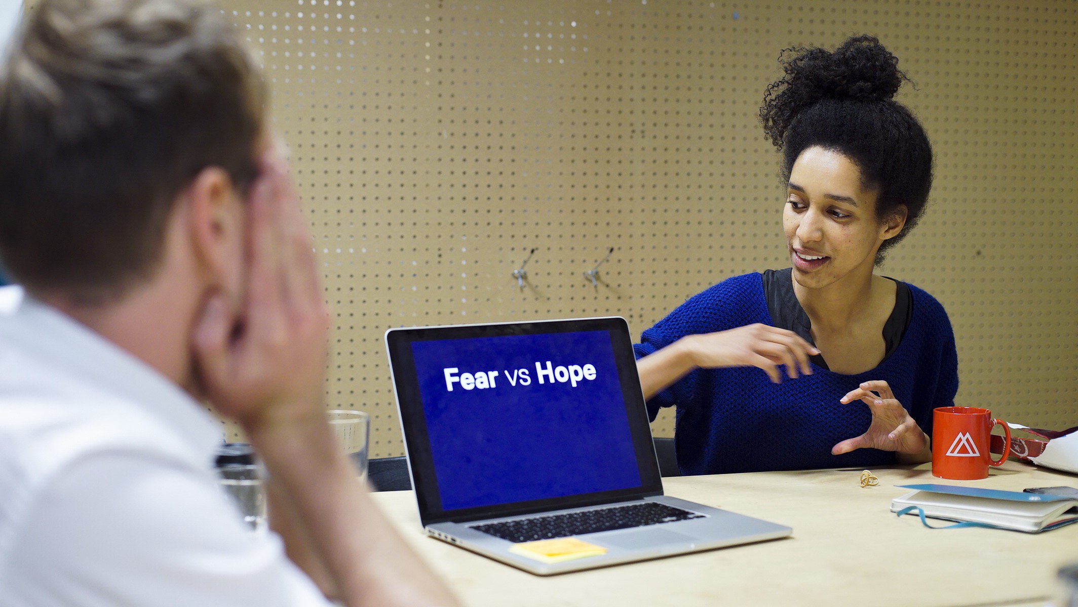 Two women having a discussion at a table. One woman is gesturing with her hand, and the other woman appears to be listening with her hand on her face. On the table, there is a laptop displaying the words "Fear vs Hope," along with a red mug and some papers.