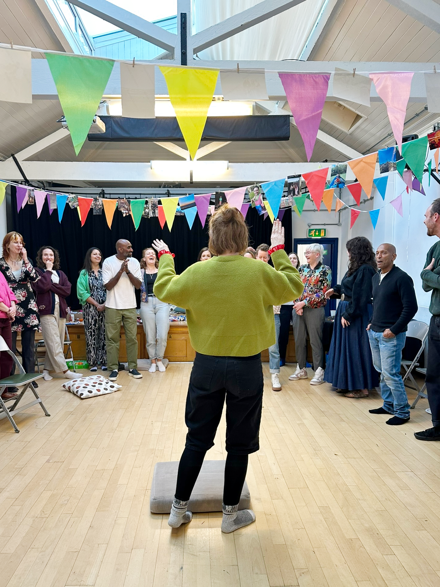 A group of people gathered in a room decorated with colorful pennant banners, with a woman standing on an elevated platform facing the group, apparently leading or performing in front of them.
