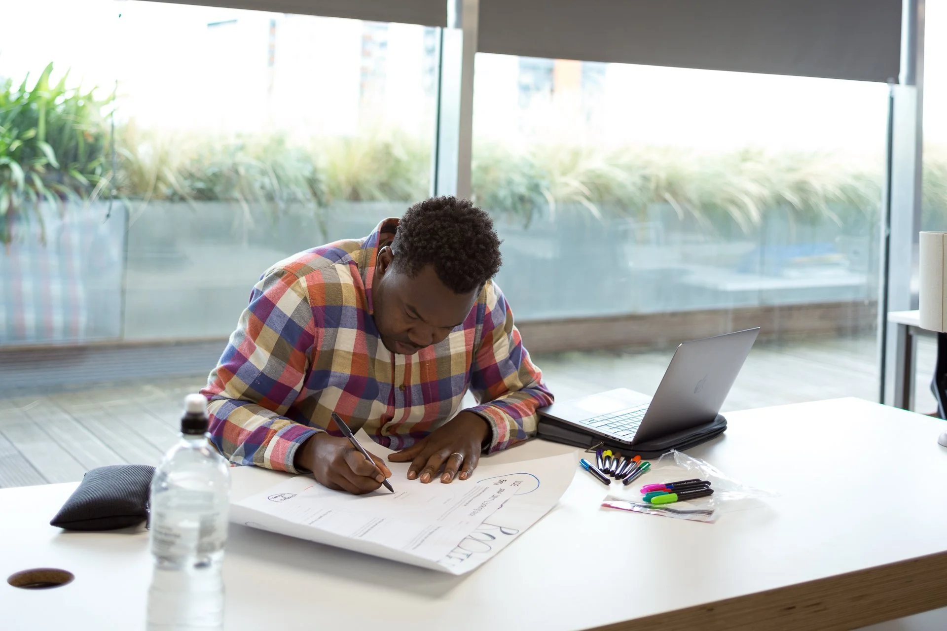 A man sitting at a desk writing on a large document with markers and a laptop nearby, in a bright room with large windows and view of plants outside.