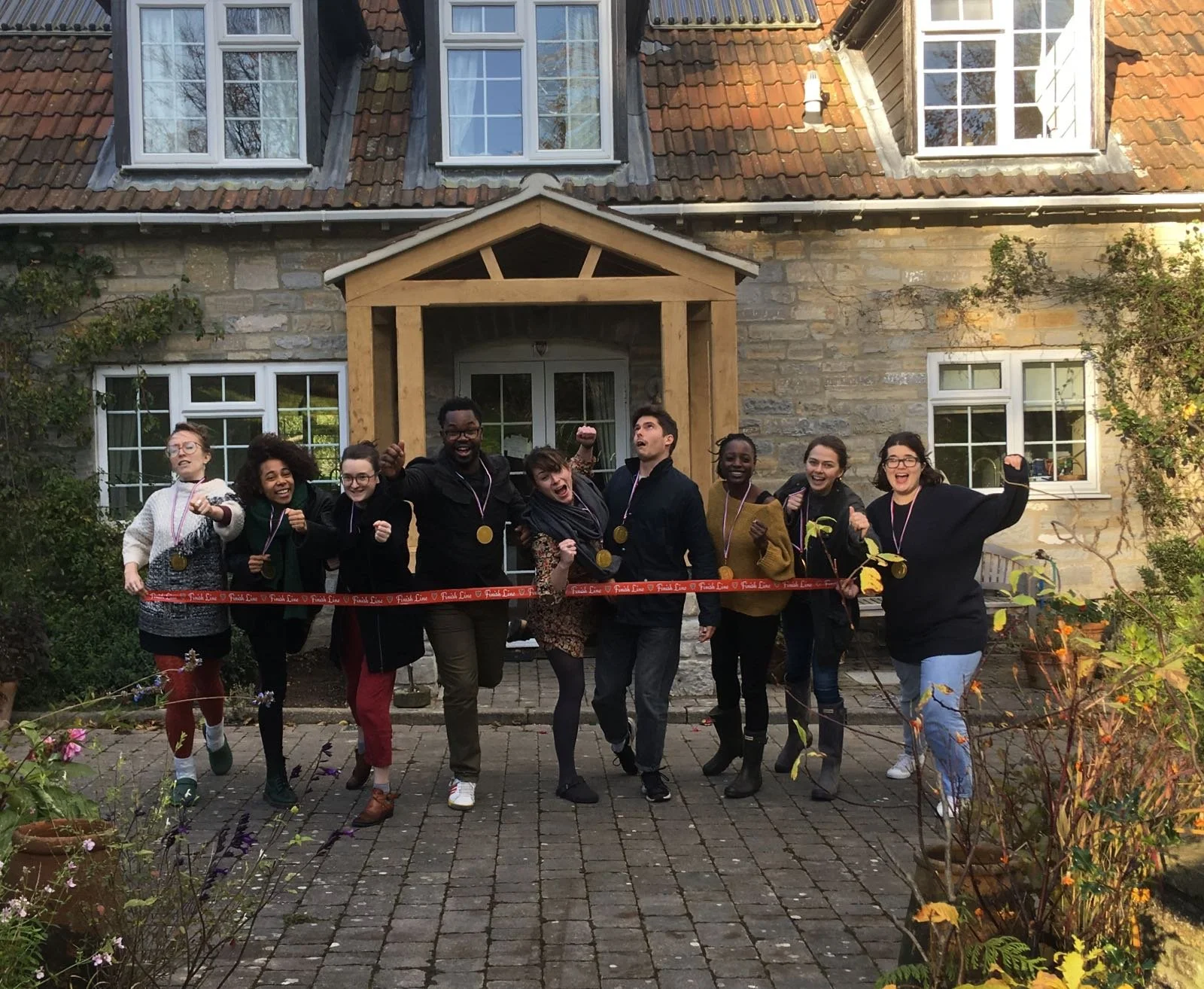 A group of ten people celebrating a ribbon-cutting ceremony outside a house with a stone facade. They are smiling, raising their fists, and wearing medals around their necks.