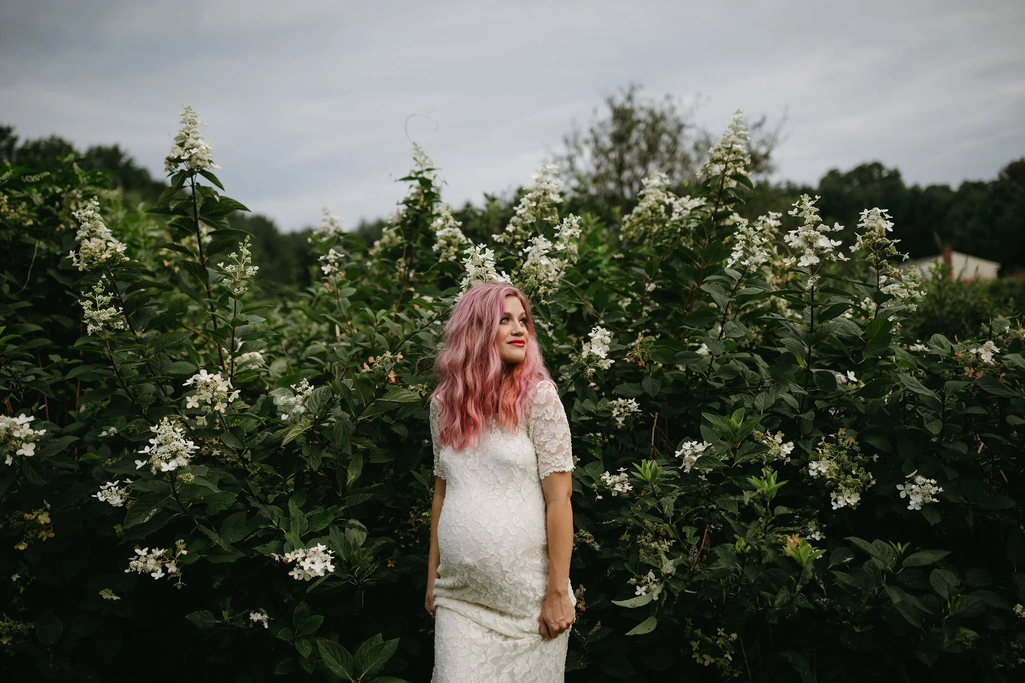 A woman with pink hair in a white lace dress standing among blooming bushes with white flowers outdoors.