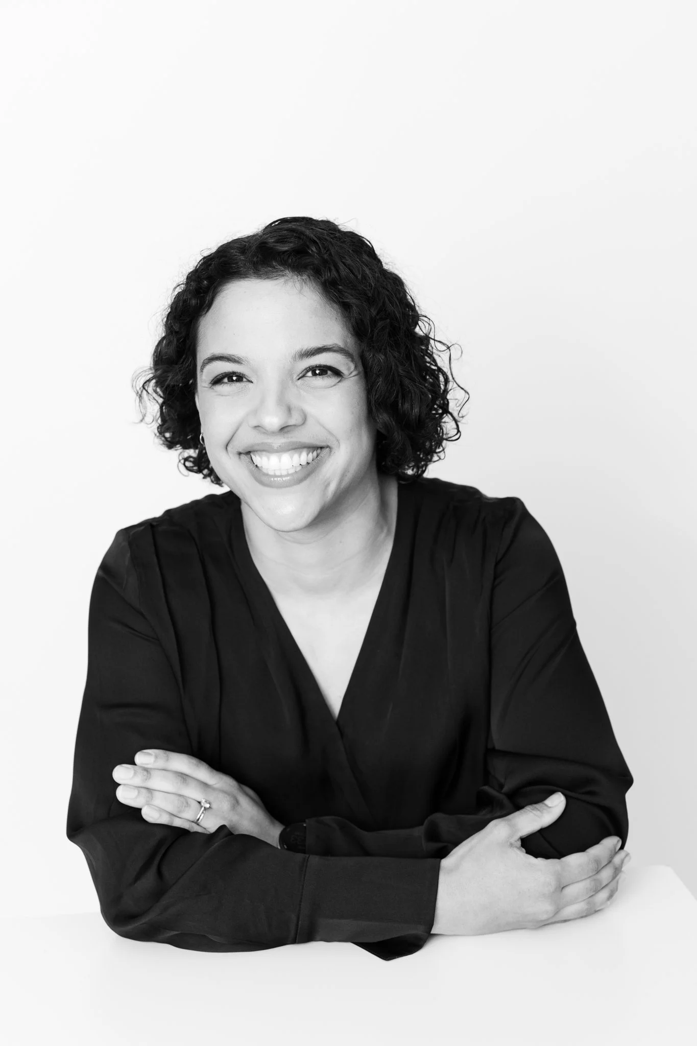 Black and white portrait of a woman with curly hair smiling, wearing a dark blouse, arms crossed on a white table against a plain white background.