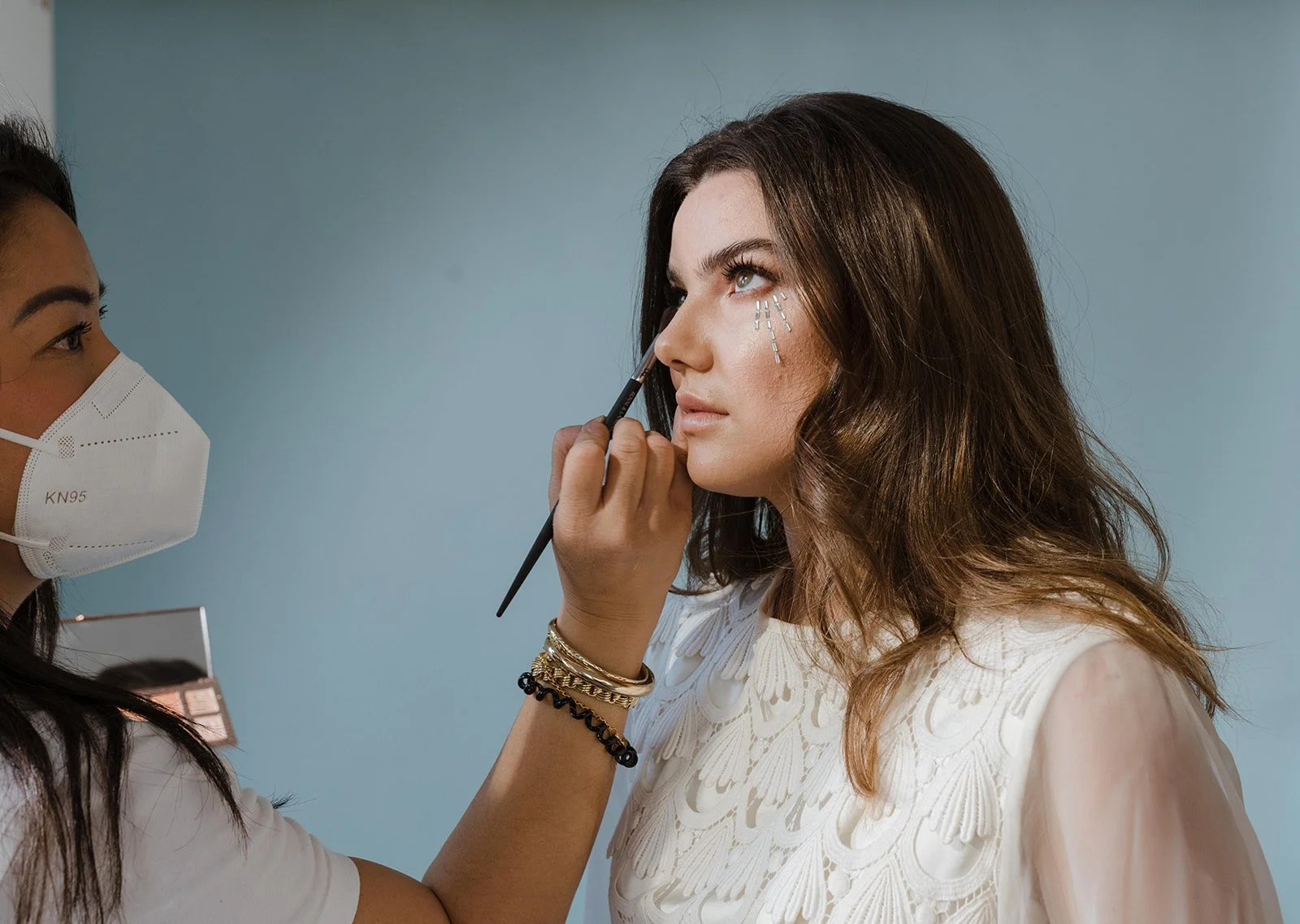 Makeup artist applies eye makeup to a woman with brown wavy hair, while wearing a white lace top, as the woman looks upward. The makeup artist wears a KN95 face mask and has bracelets on her wrist.