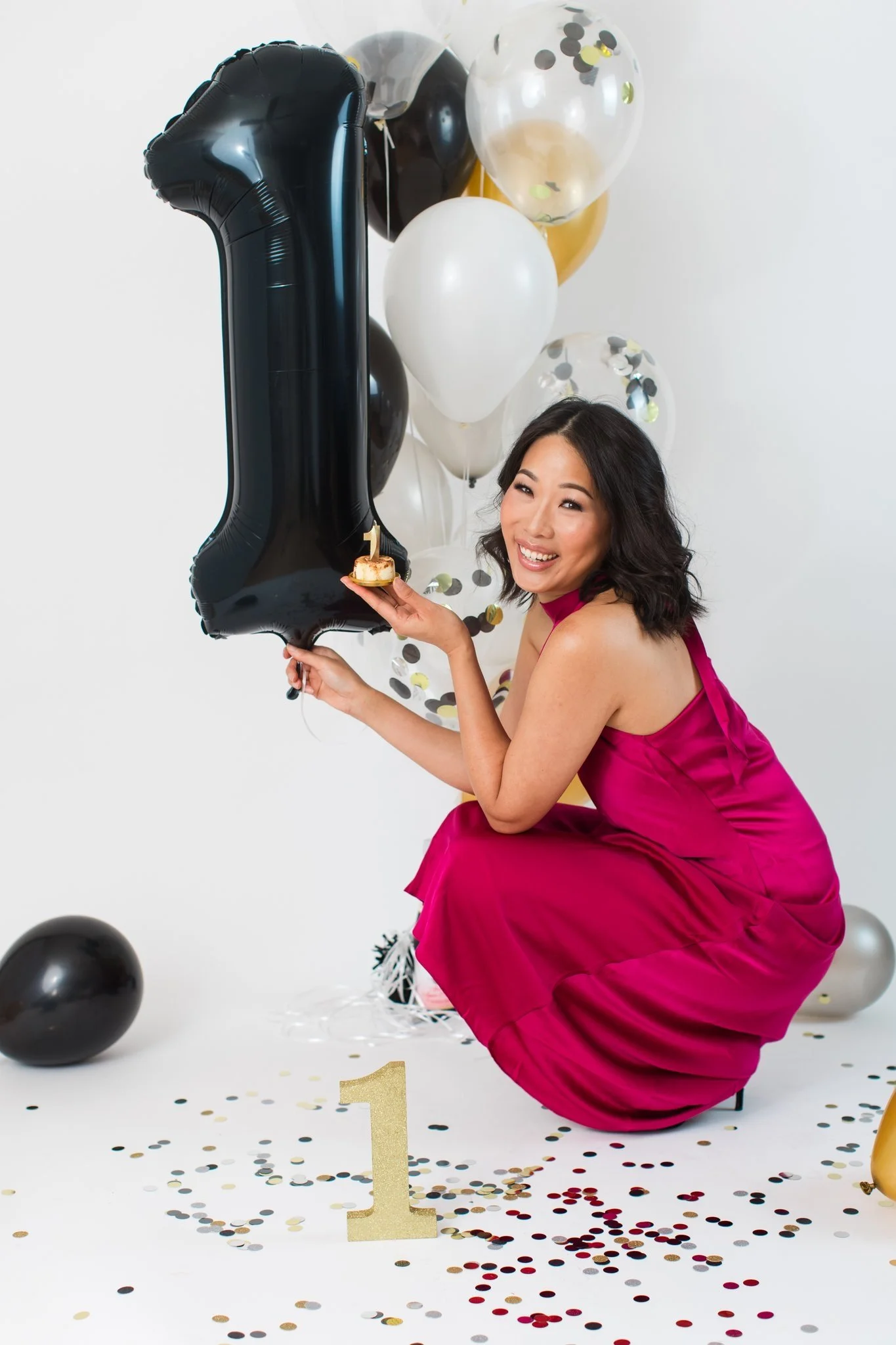 A woman in a pink dress kneeling on the floor, holding a cake with a number one candle, surrounded by black, white, gold, and transparent balloons, and confetti, celebrating her first birthday.