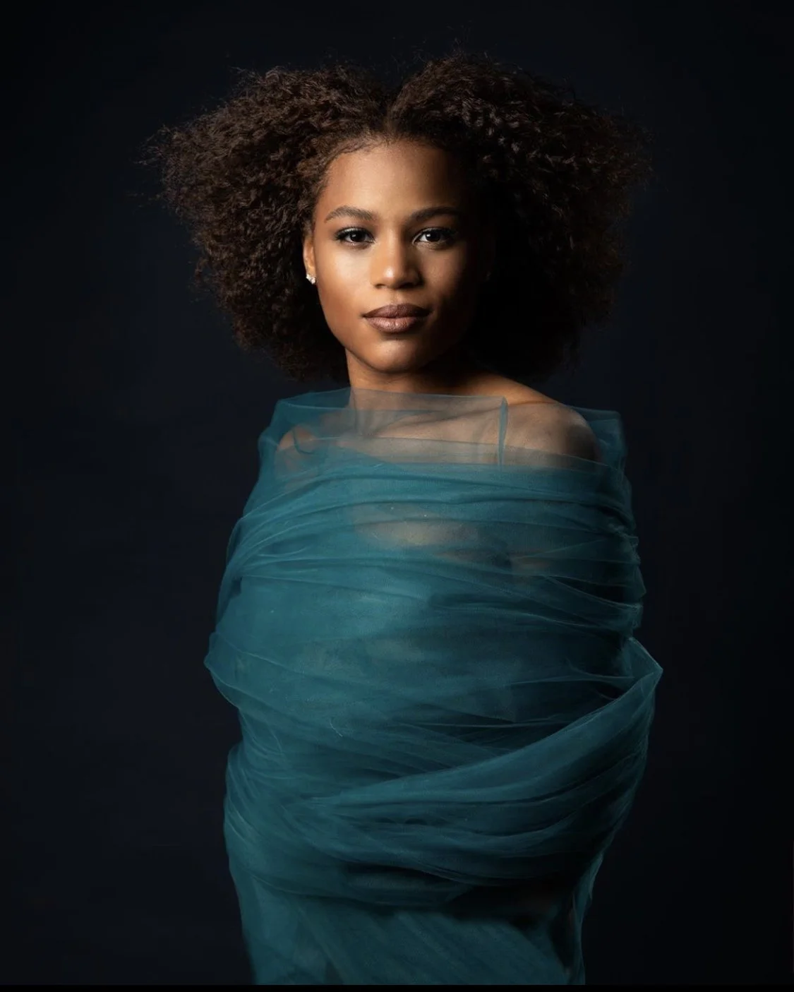 Portrait of a woman with curly hair wrapped in sheer teal fabric against a dark background.