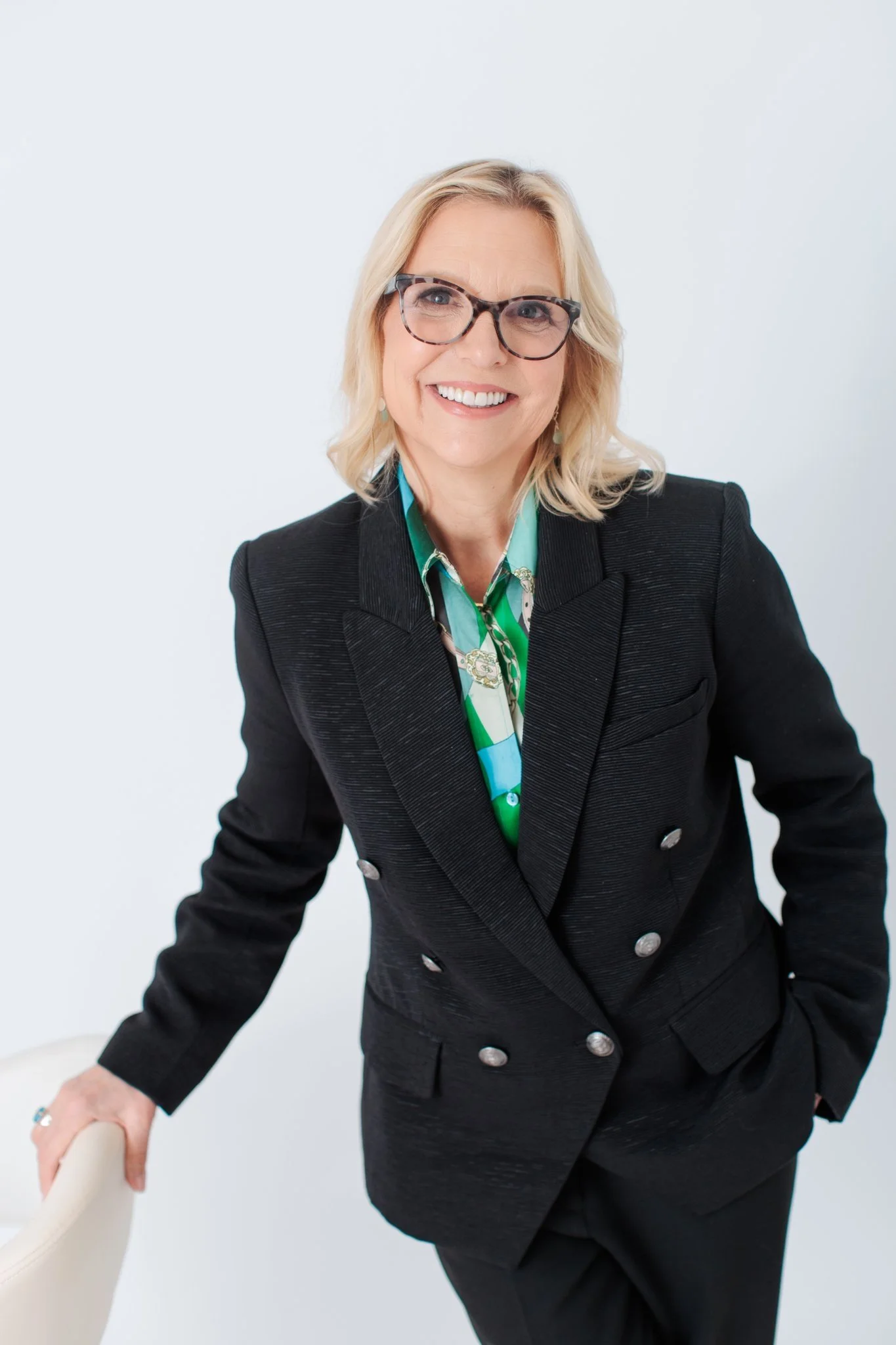 A woman with blonde hair and glasses smiling, wearing a black blazer over a colorful blouse, standing and leaning on the back of a cream-colored chair against a plain white background.
