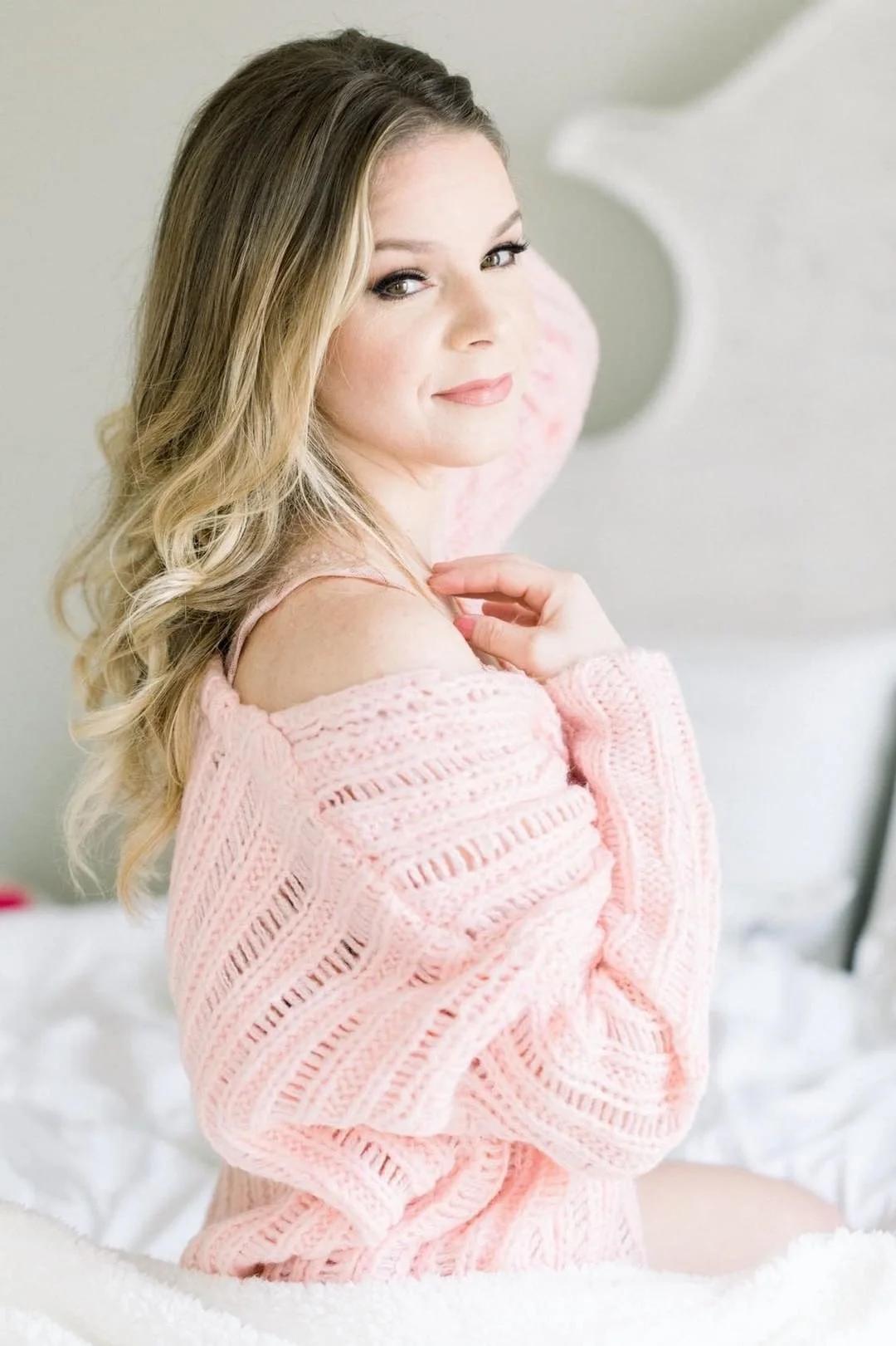 A woman with long blonde hair, wearing a pink knit sweater, sitting on a bed in a softly lit room, looking at the camera with a gentle smile.