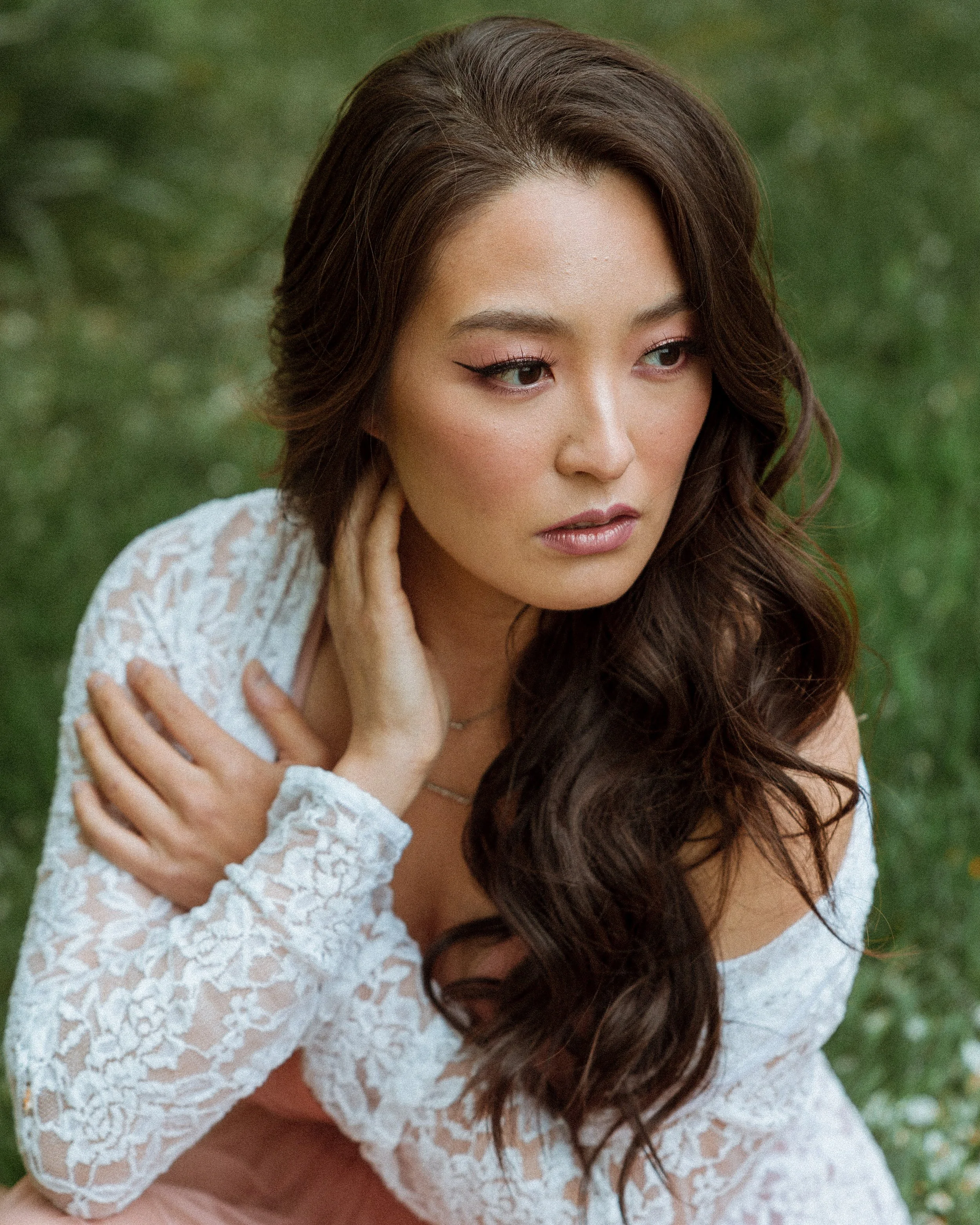 A woman with long wavy brown hair in a white lace top, sitting outdoors on green grass, looking contemplative.
