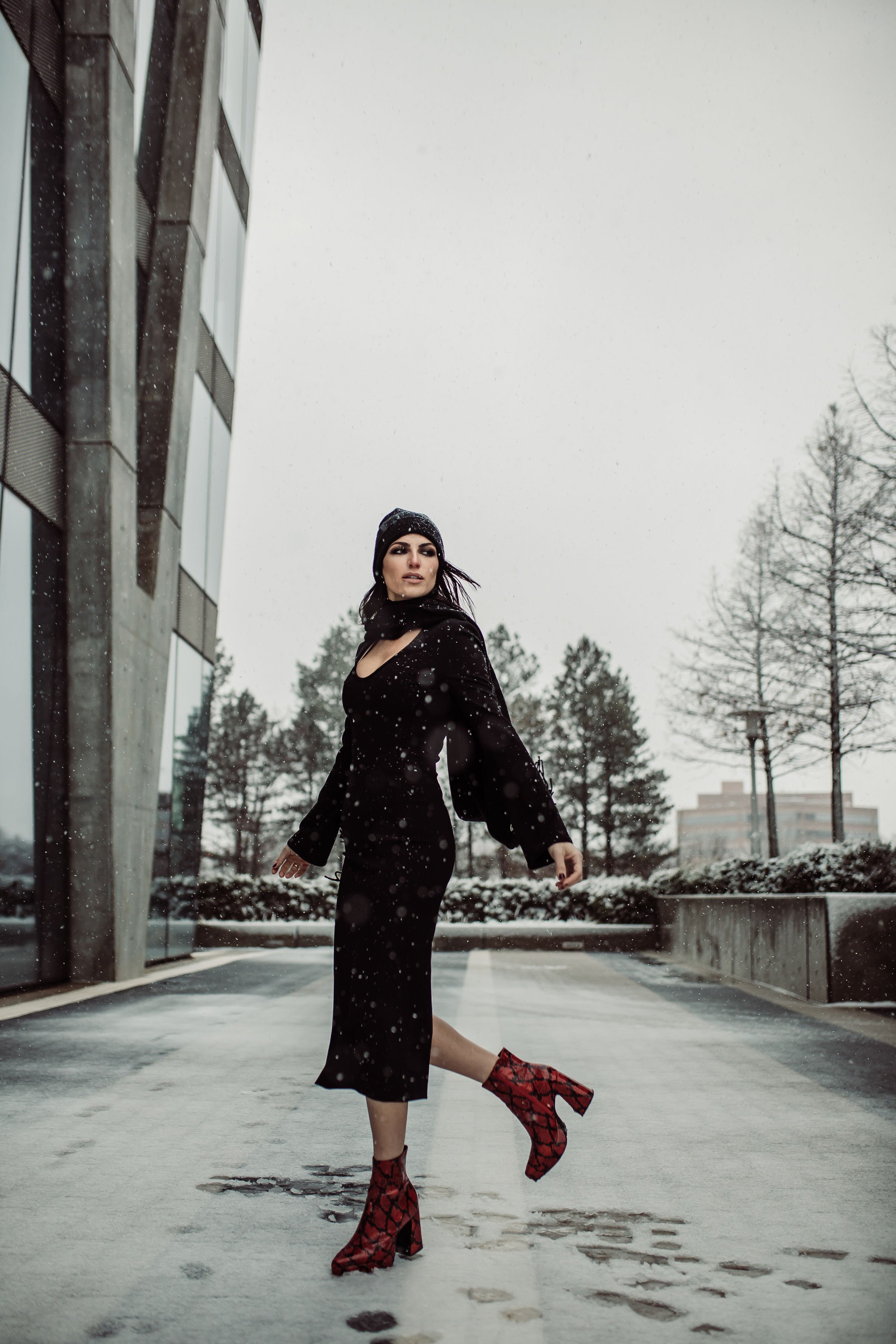 A woman in a black dress and red snake-skin patterned ankle boots walking outside on a snowy day near modern buildings and leafless trees.
