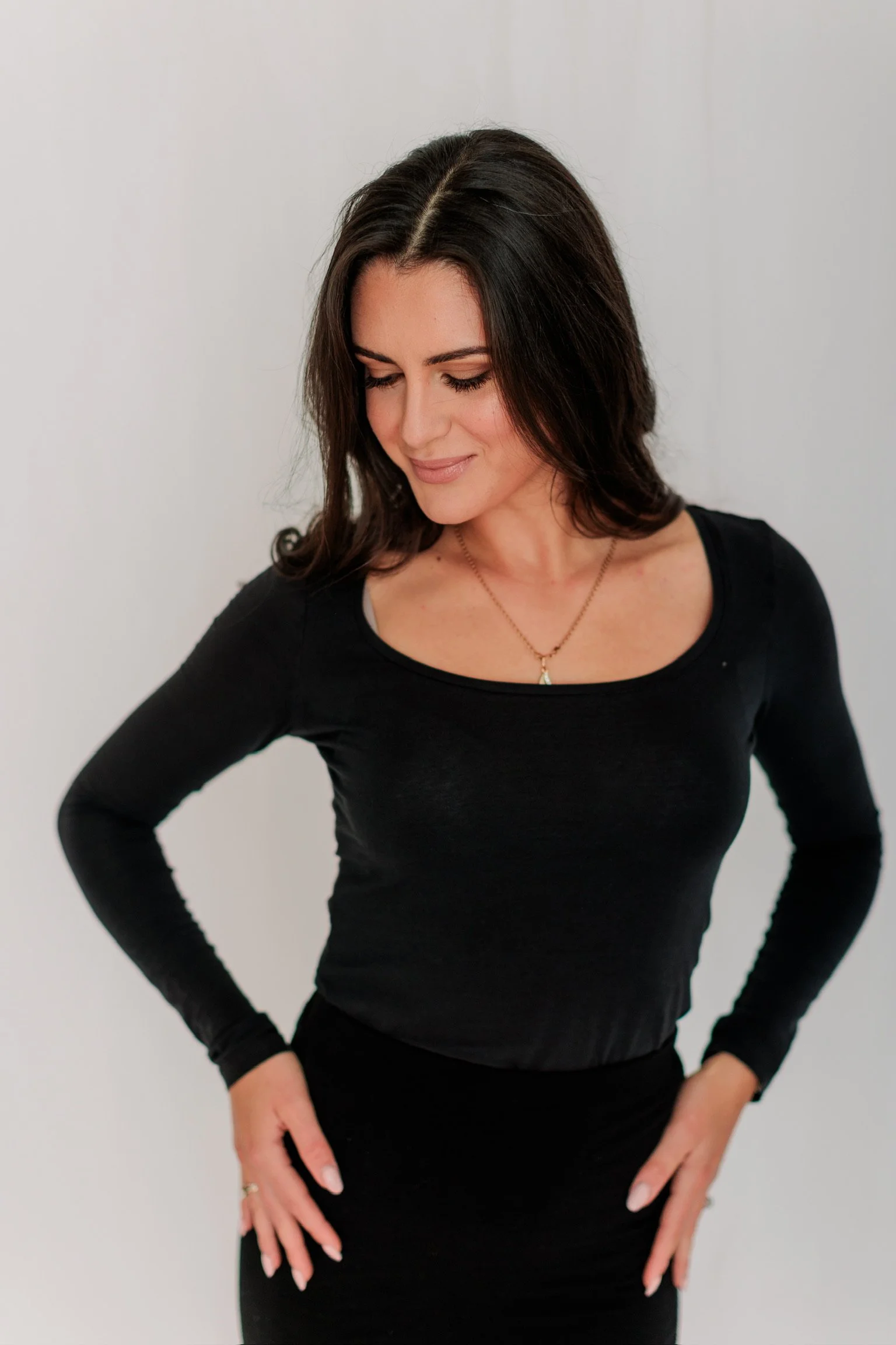 A woman with dark brown hair wearing a black long-sleeve shirt and black skirt, looking down with her hands on her hips, standing against a plain wall.