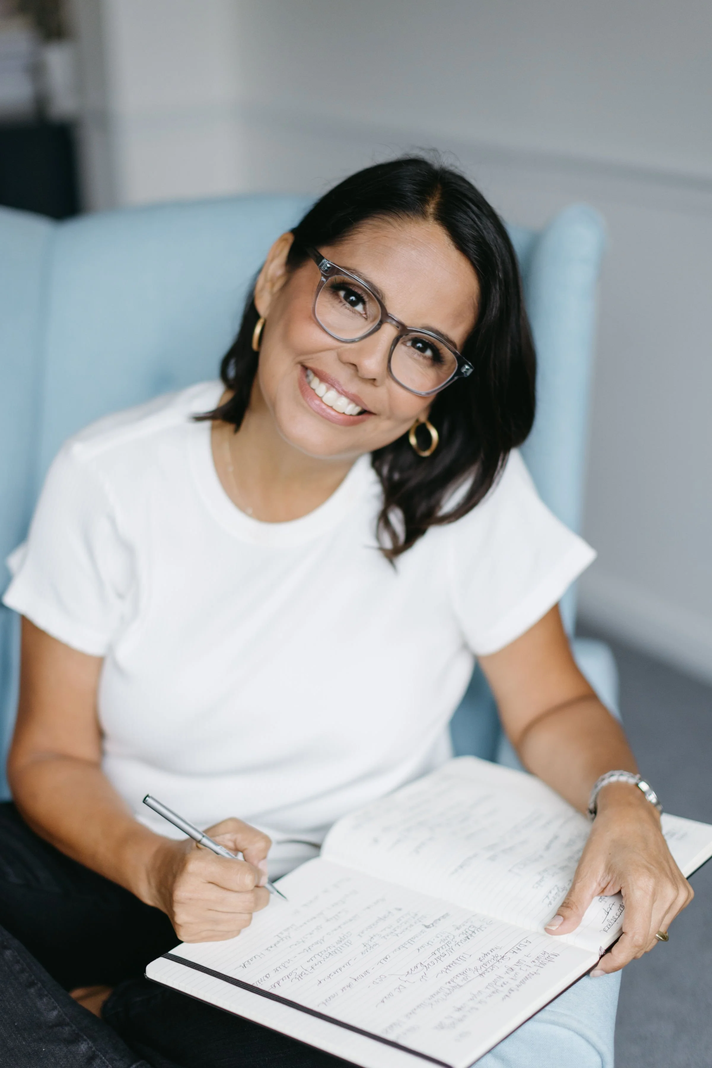 A woman with glasses, wearing a white shirt and gold hoop earrings, sitting on a light blue armchair, smiling, and writing in a notebook.