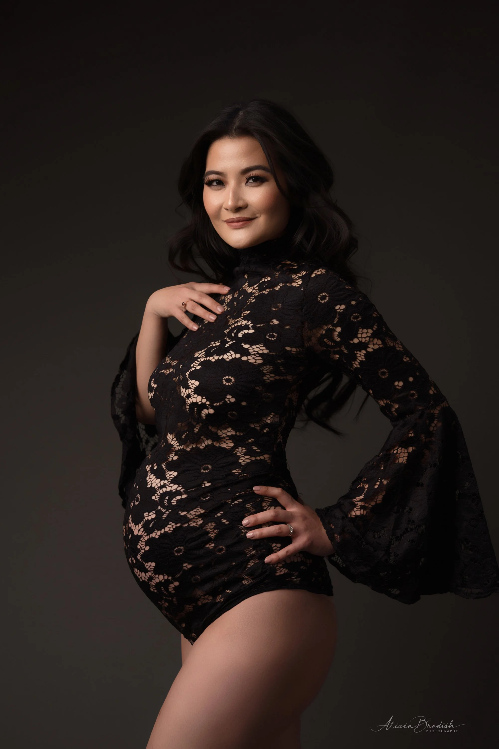 A woman in a black lace dress with sheer bell sleeves posing with hand on hip and smiling, dark background.