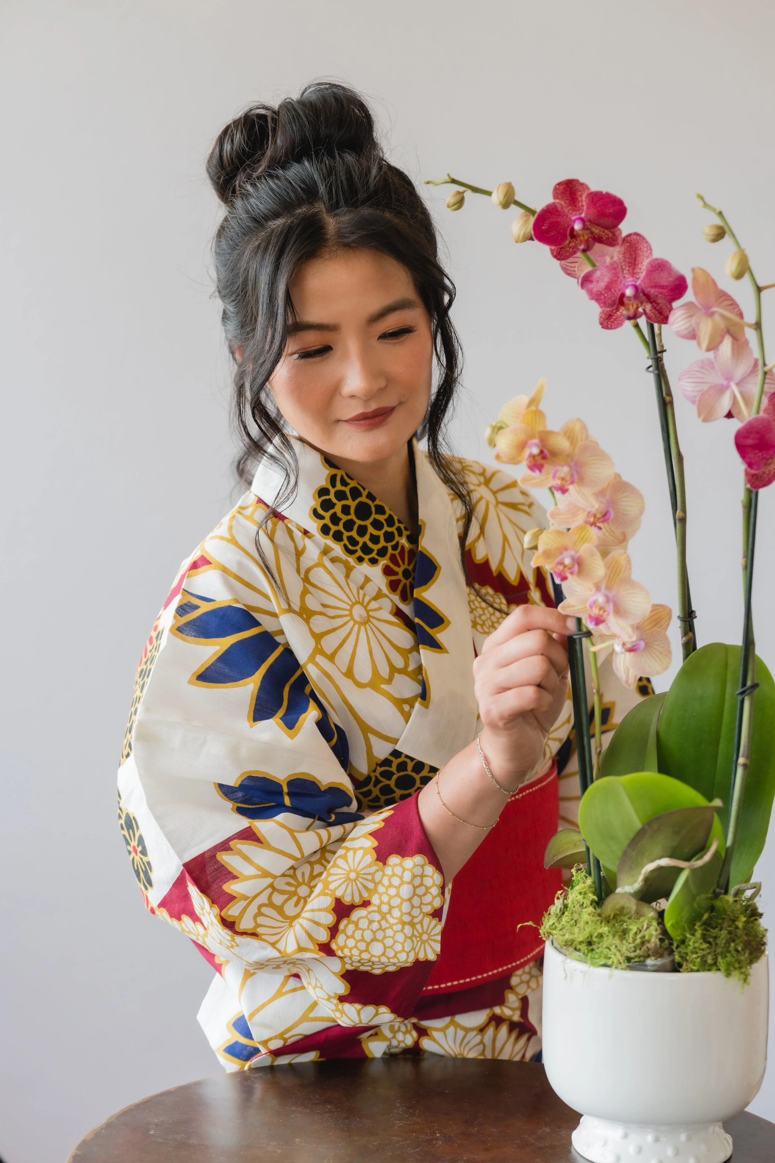 A woman with dark hair styled in a bun is arranging pink and cream orchids in a white pot.