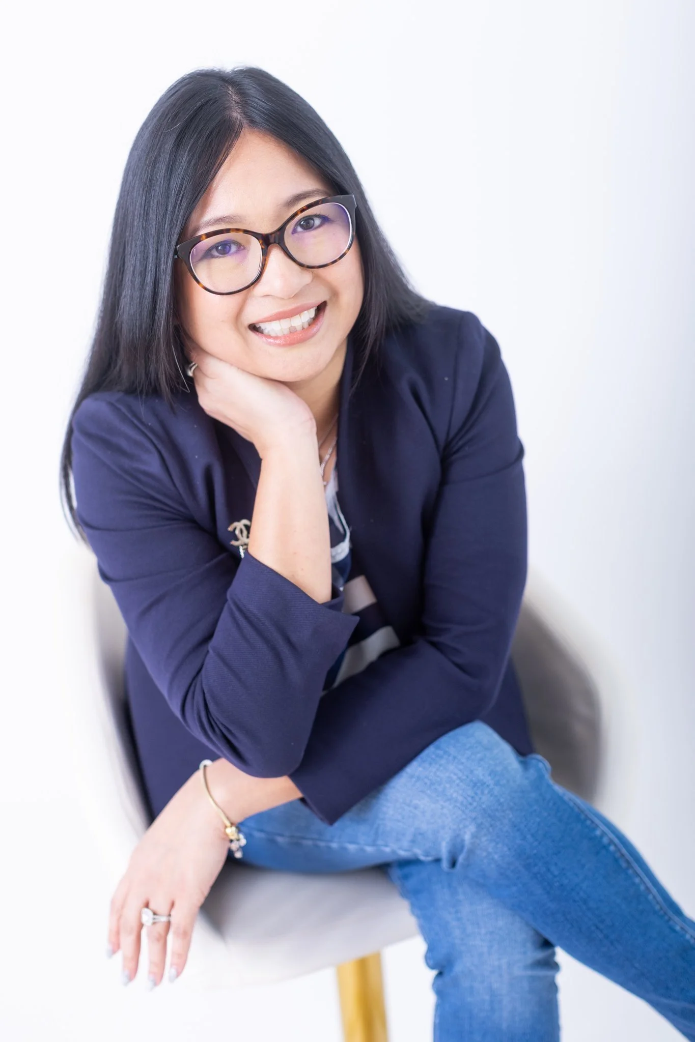 A woman with long black hair and glasses, wearing a navy jacket and jeans, sitting on a chair and smiling at the camera.