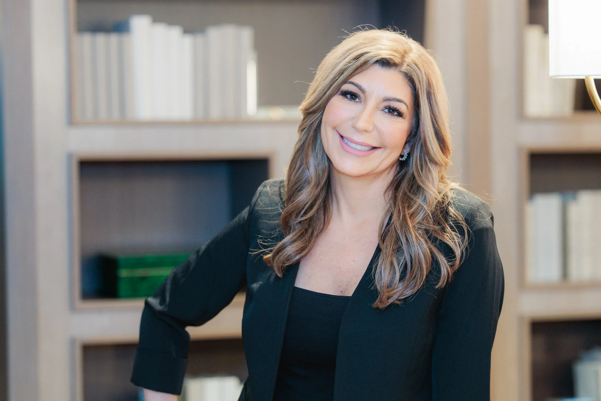A woman with long, wavy brown hair and light skin smiling while leaning forward slightly in front of a bookshelf filled with books. She is wearing a black blazer and earrings.