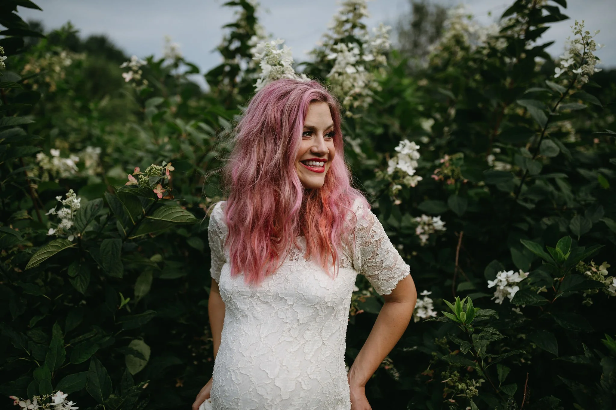 A woman with pink, wavy hair smiling and posing outdoors among green foliage and white flowers, wearing a white lace dress.