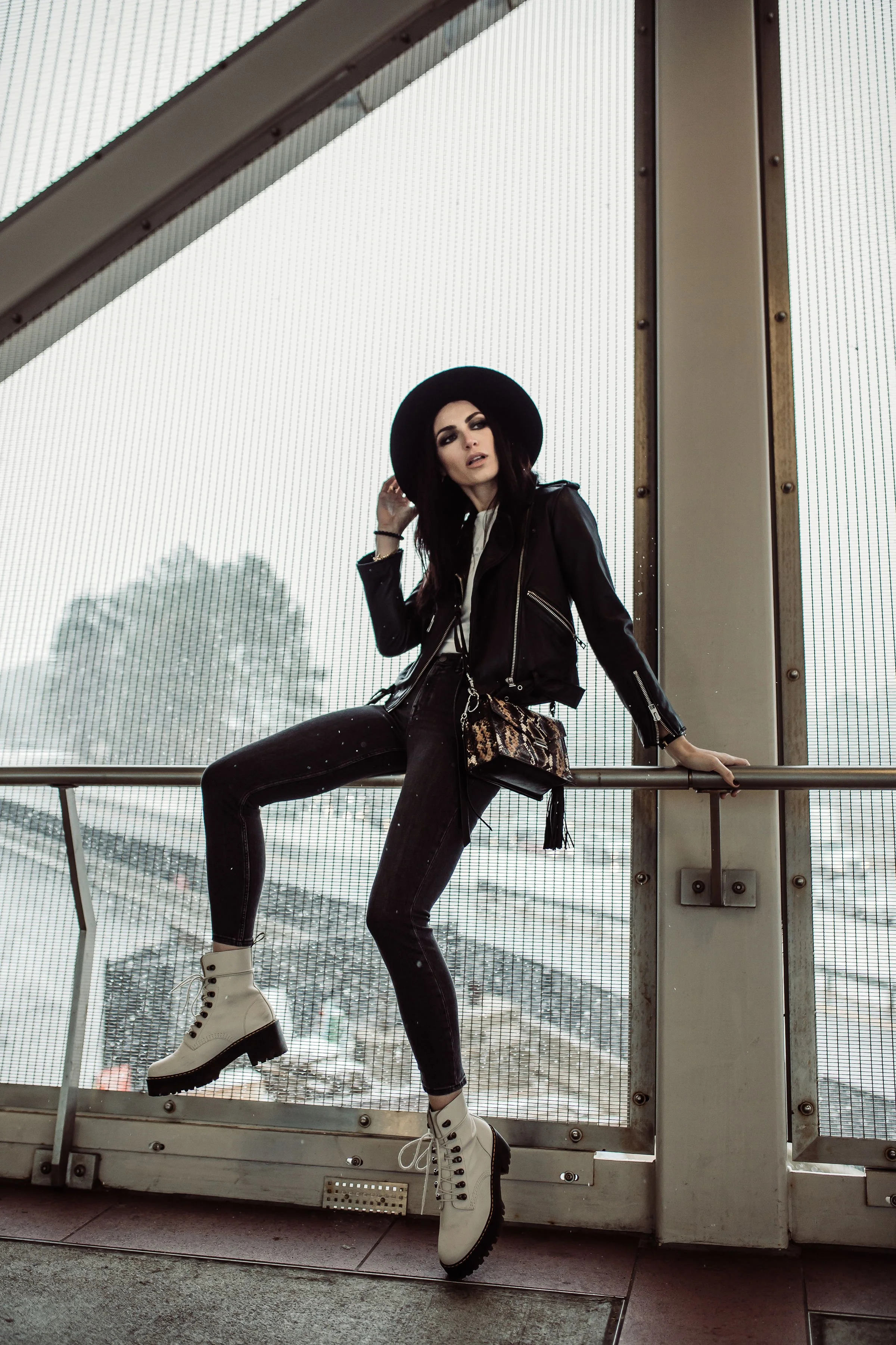 Fashionably dressed woman in black leather jacket, black jeans, white combat boots, and black hat, sitting on railing inside a building with a large window background.