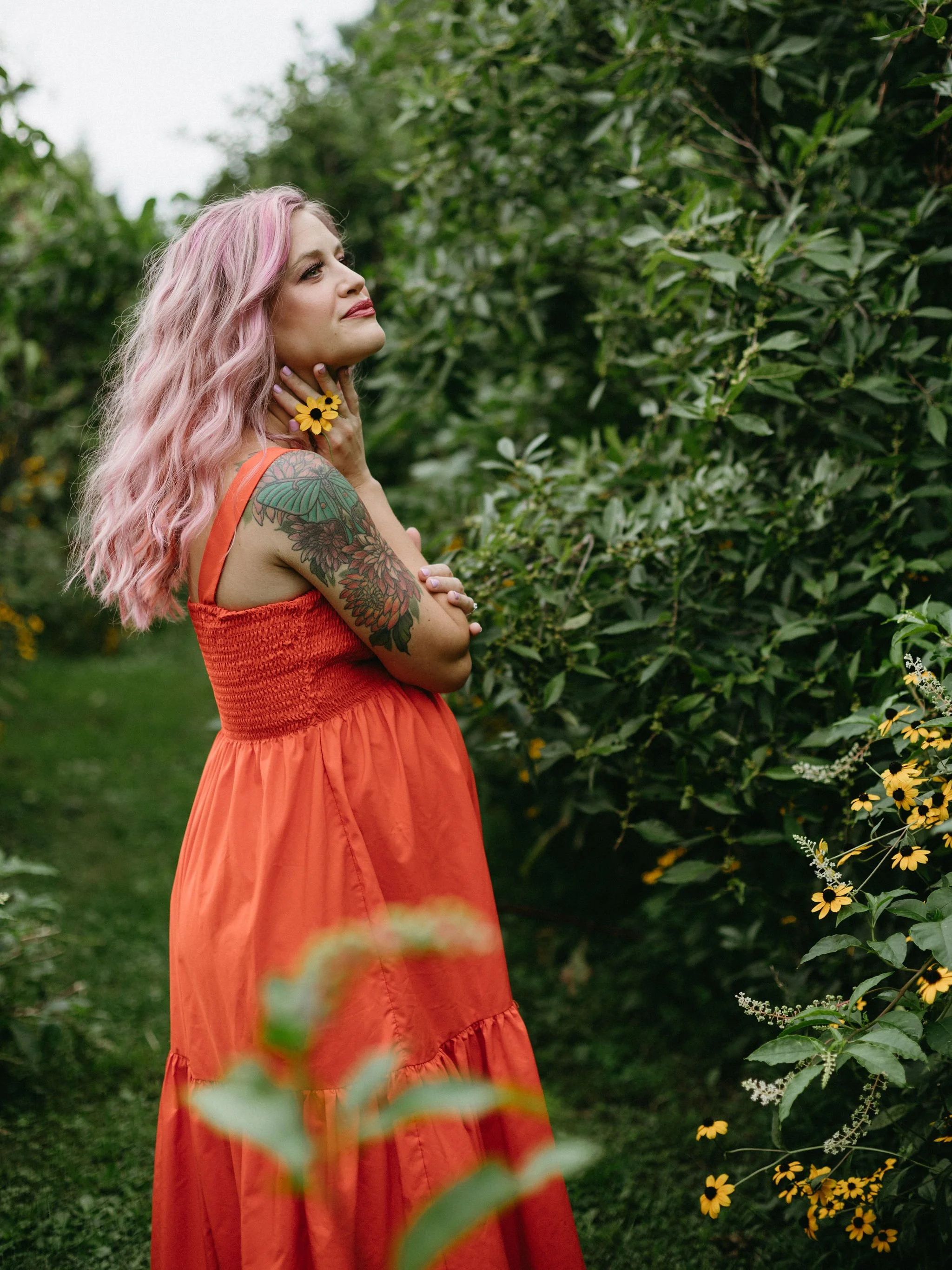 A woman with pink hair wearing an orange dress with floral tattoos on her arm, standing in a lush garden with yellow flowers.