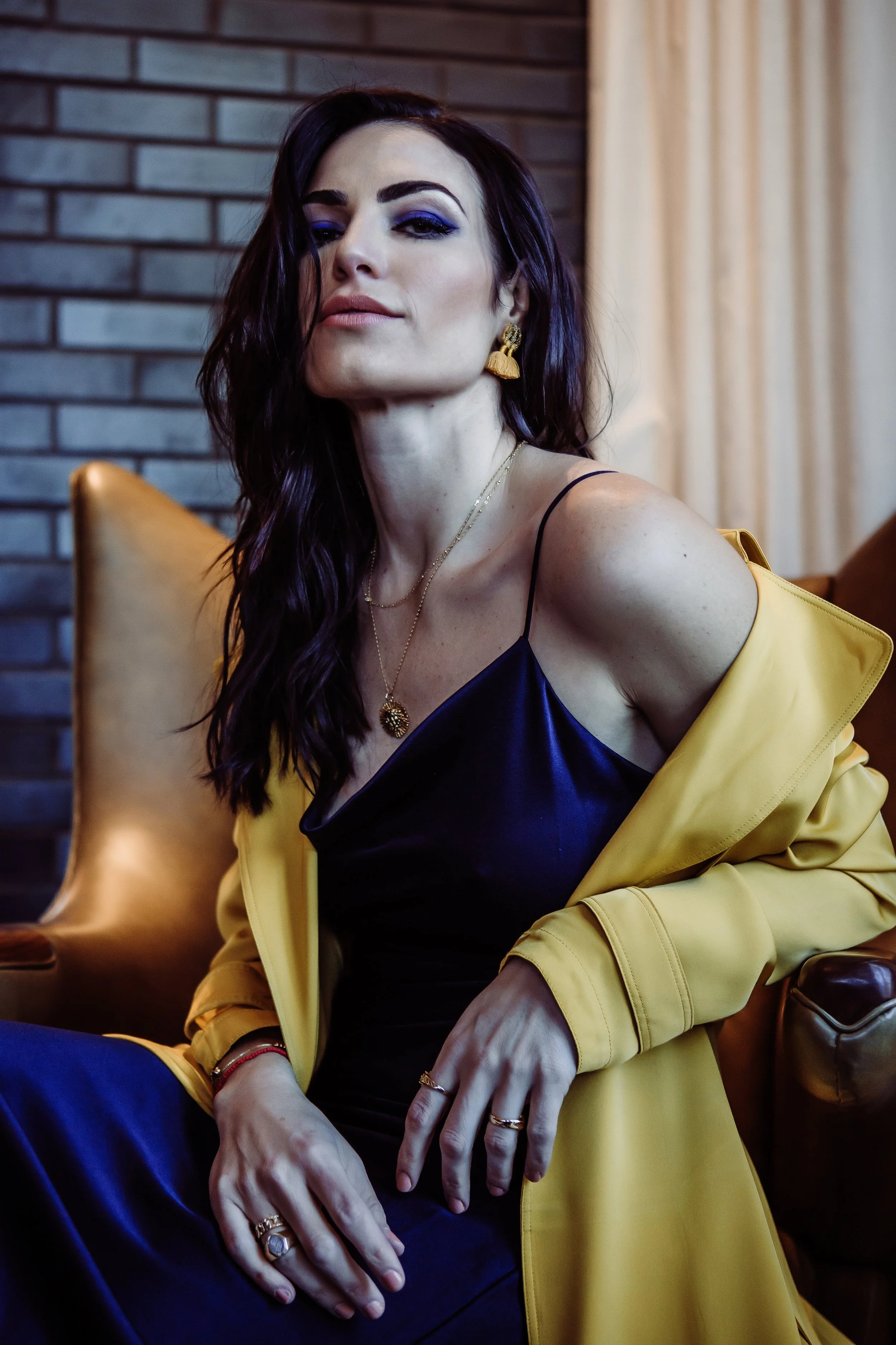 A woman with dark wavy hair and blue eyeshadow sitting on a leather armchair against a brick wall, wearing a black satin dress and a yellow jacket, accessorized with gold jewelry.