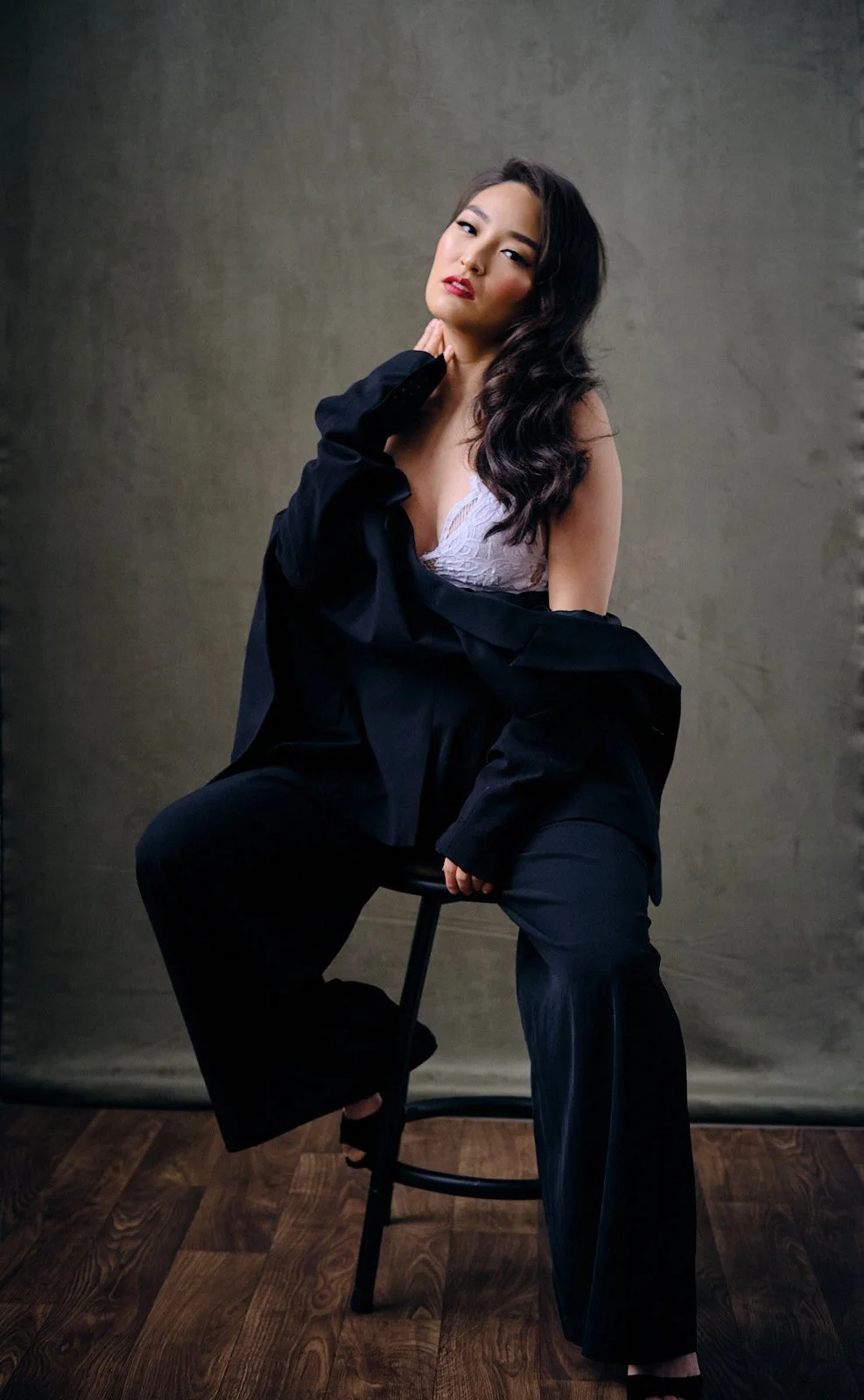 A woman wearing a black jacket draped over her shoulders, white lace top, and black pants, sitting on a tall stool against a plain, textured background with wooden floor, posing with her hand touching her neck and looking at the camera.