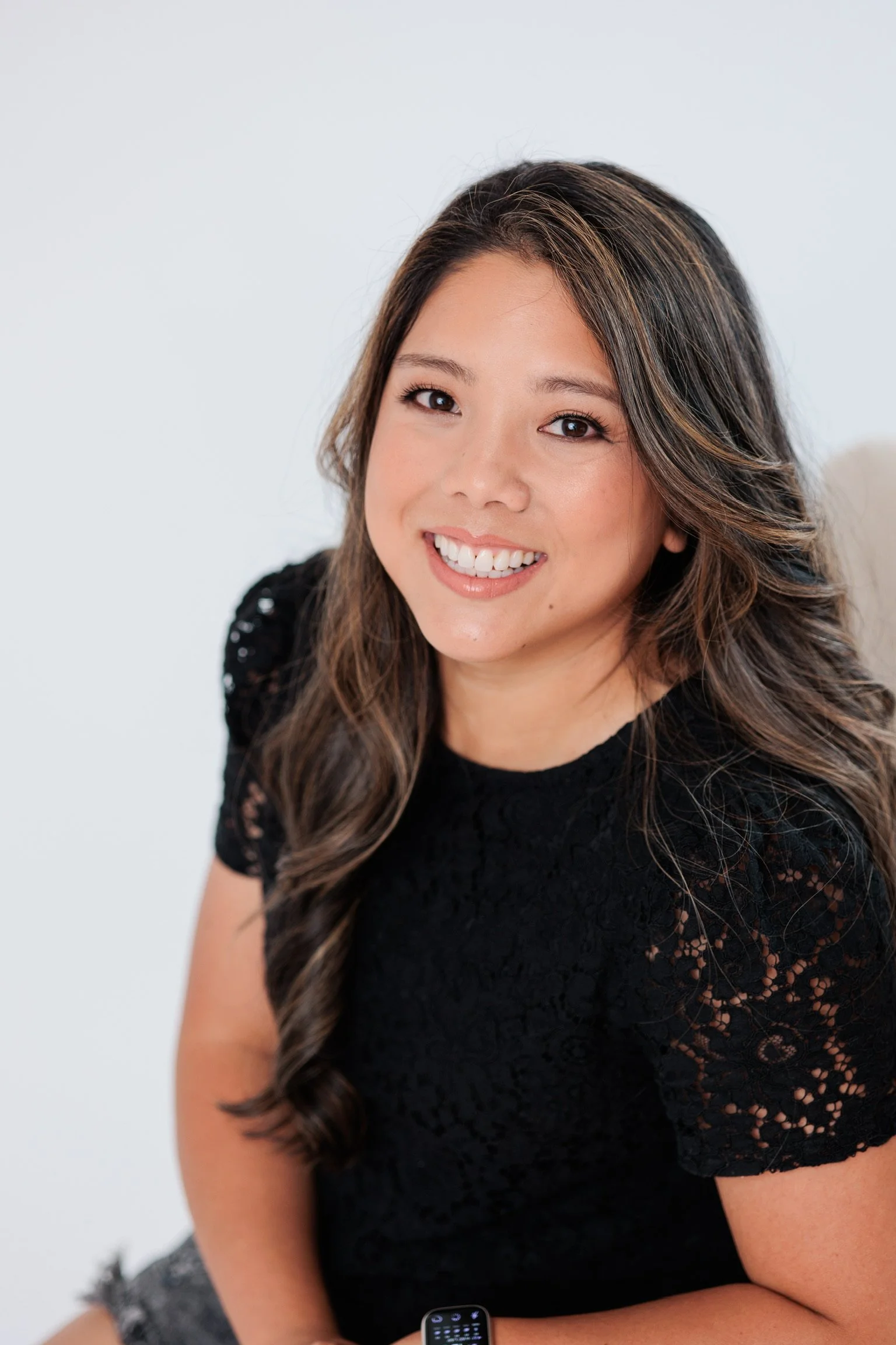 Portrait of a smiling woman with long, wavy dark hair wearing a black lace top, sitting against a plain white background.